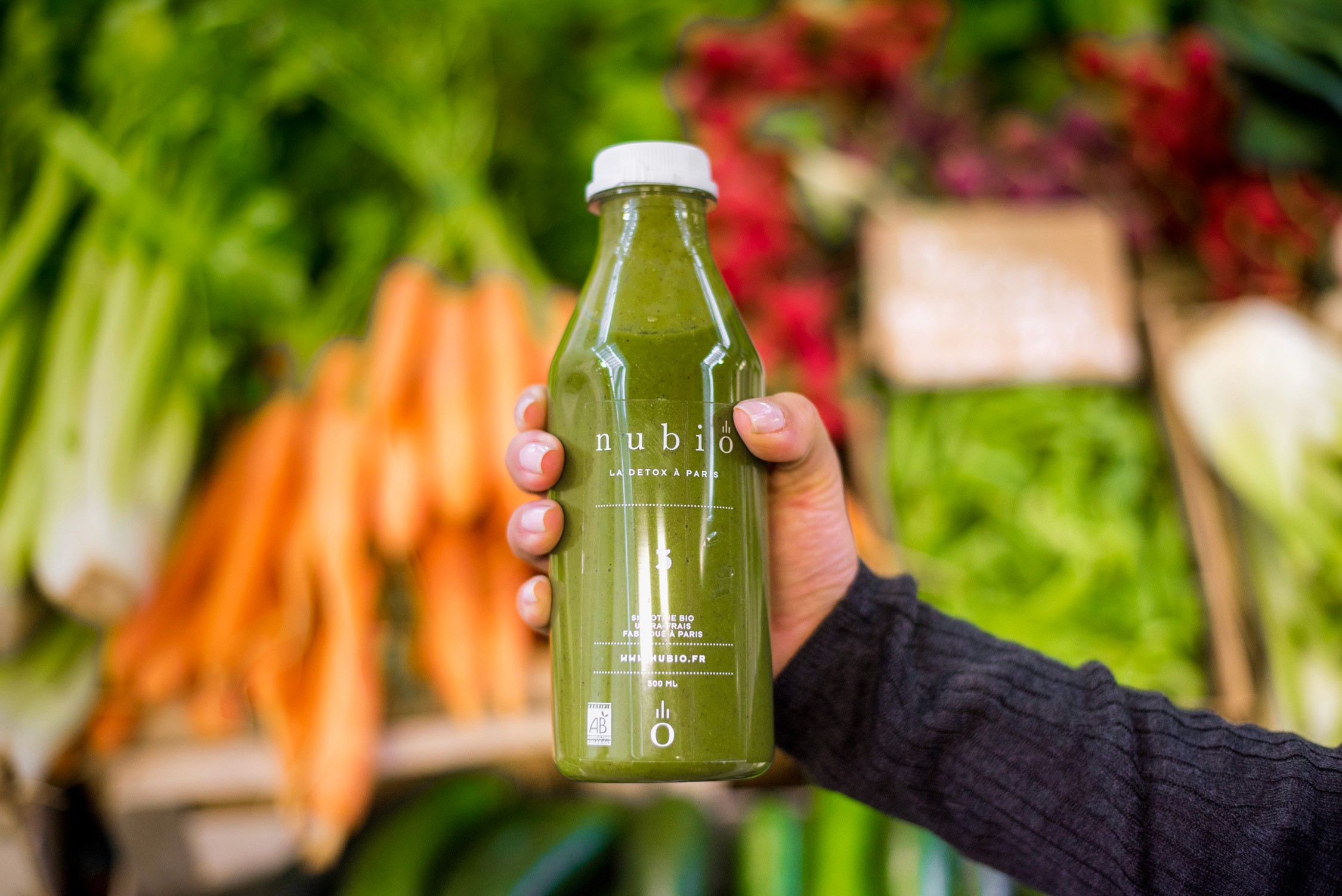 Vibrant green smoothie bottle rests amidst colorful fresh produce, suggesting a healthy market visit bathed in natural daylight.