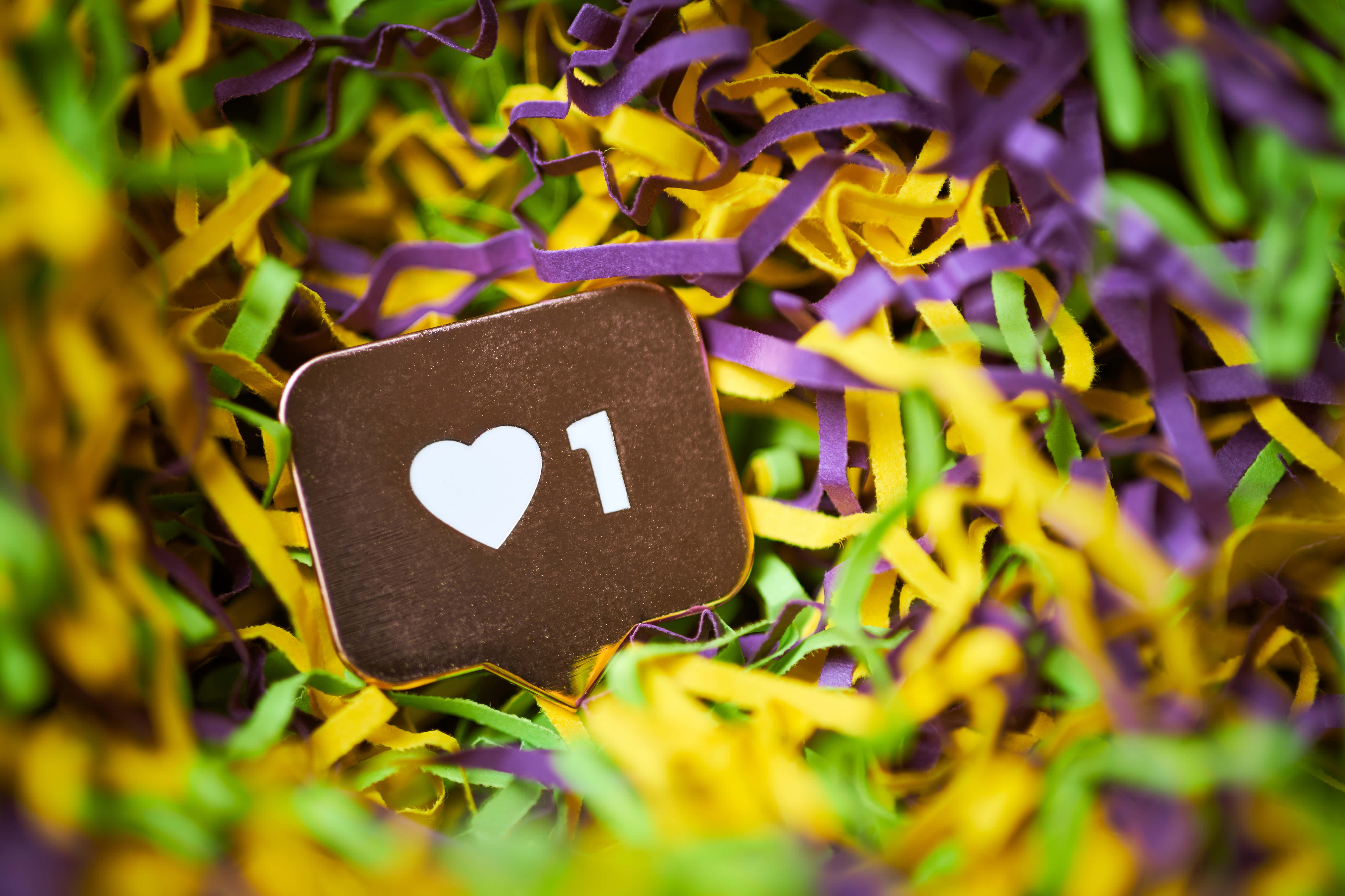 A close-up, selective focus photo of a social media 'like' icon with one heart, resting in a pile of vibrant purple, yellow, and green shredded paper, evoking a celebratory mood.