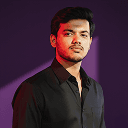 A man wearing a black shirt poses in front of a bold purple wall, highlighting his attire against the colorful background.