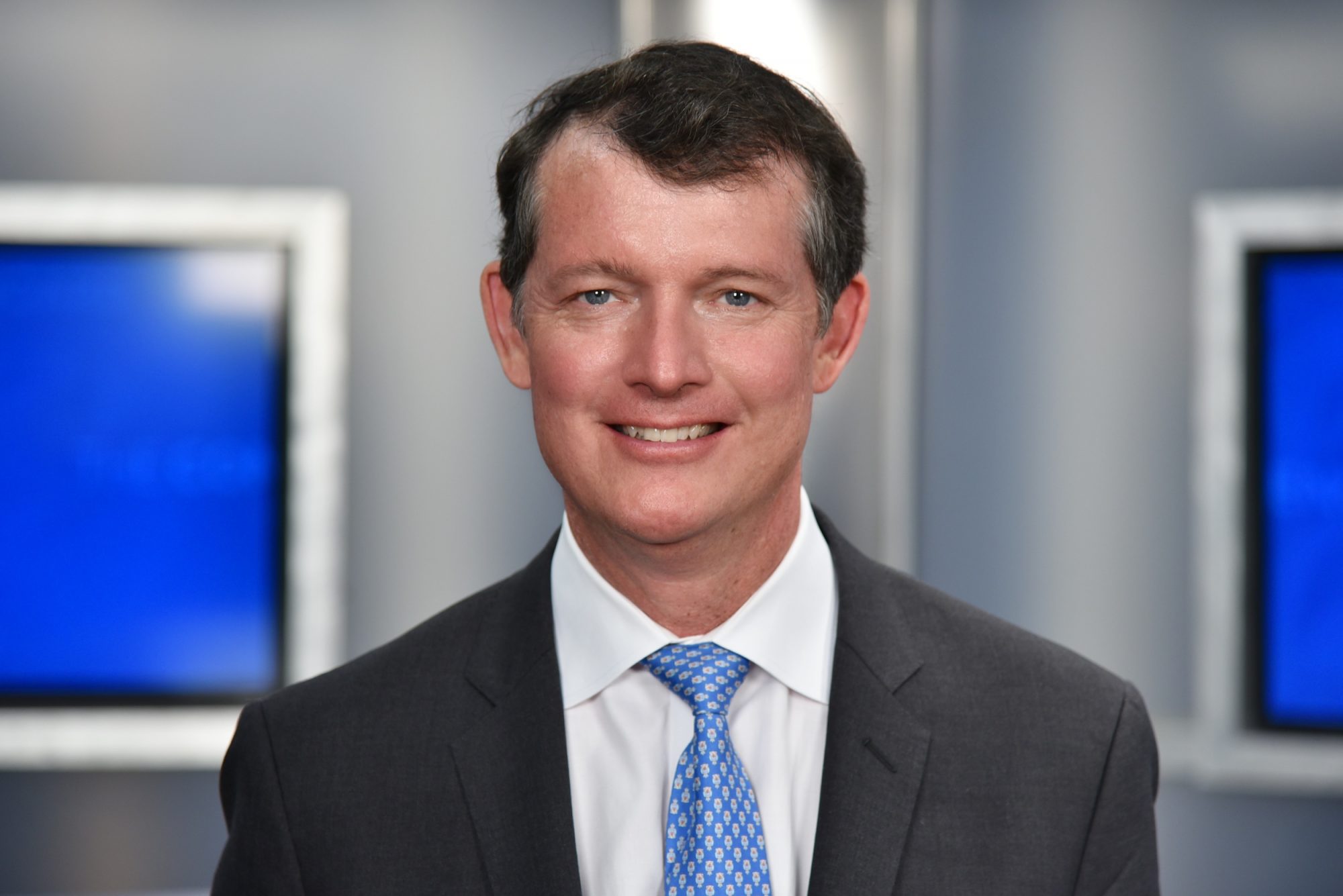 A smiling man in a suit and tie stands in front of blue backdrop screens.