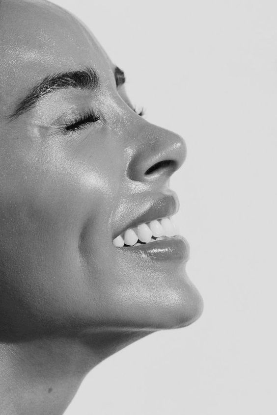 A cozy and inviting spa room with a neatly made bed, soft lighting, and warm wooden tones. The scene is partially framed by green plants, adding a natural and soothing touch.