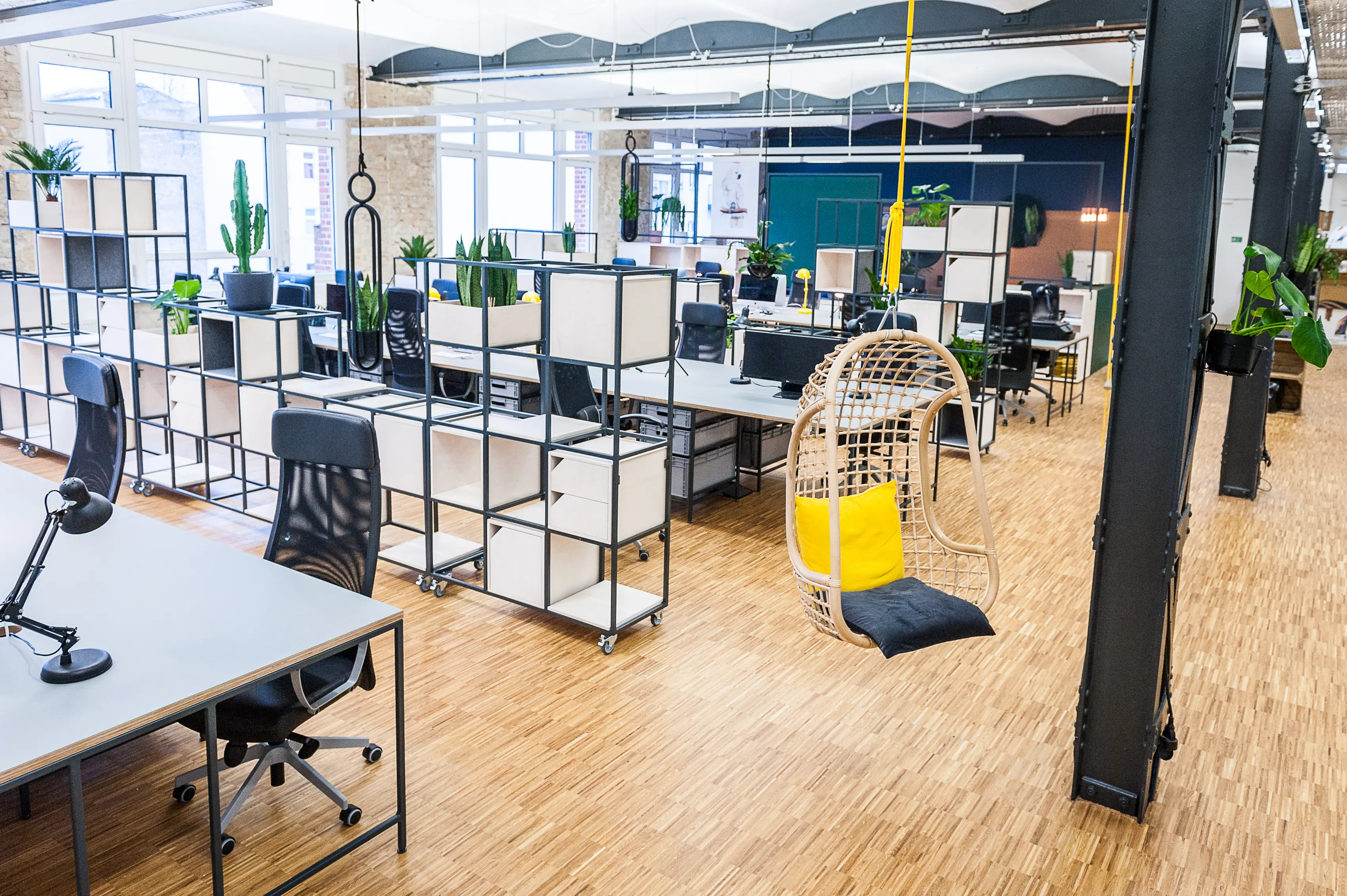 Open-plan office at INTERMATE x TRUEMATES with rows of desks divided by modular shelving units, black office chairs, a hanging rattan egg chair with yellow cushion and a vaulted white ceiling.