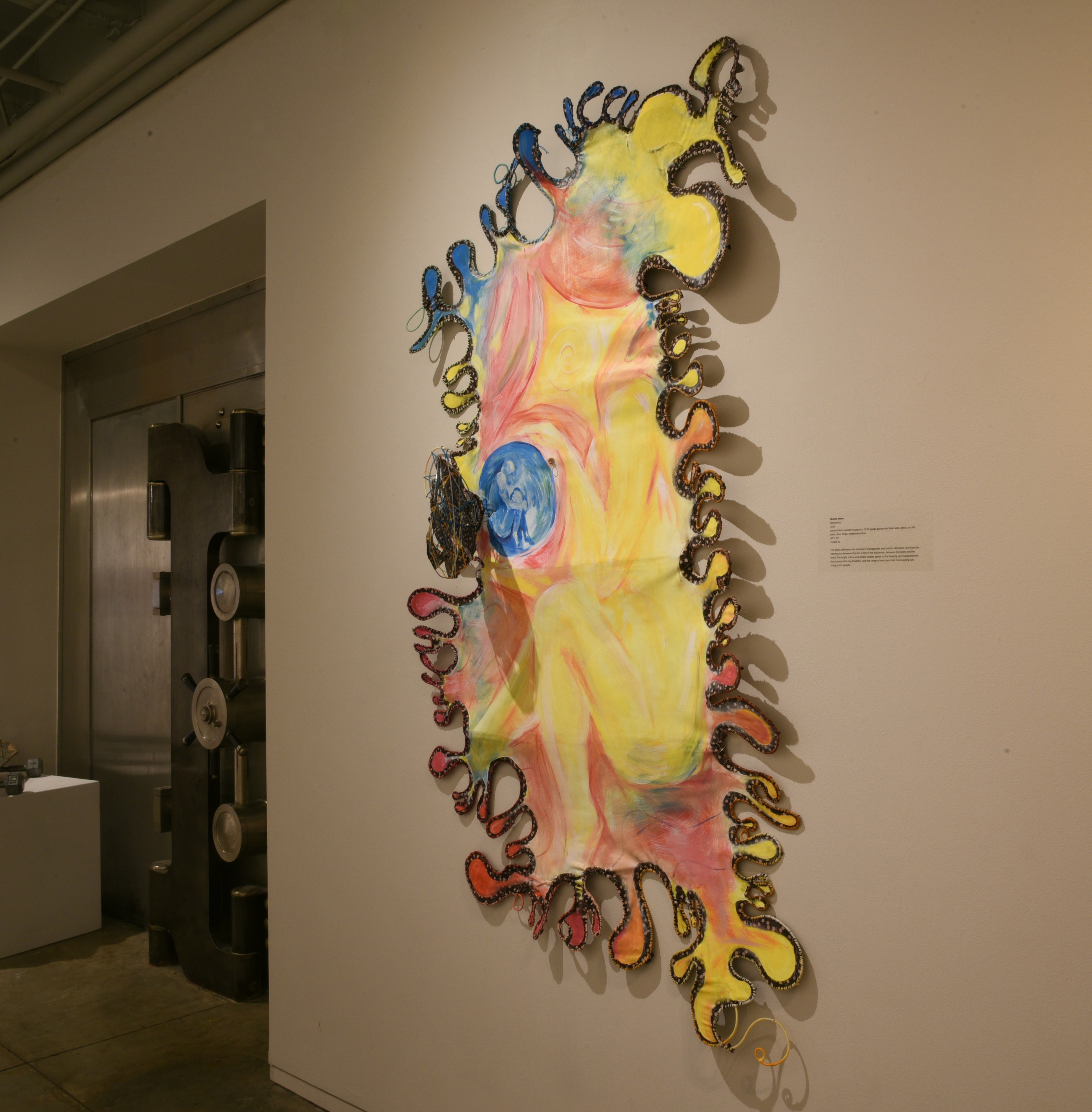 A sculpture hung on the wall at an art exhibition. It's light pink, yellow and blue.