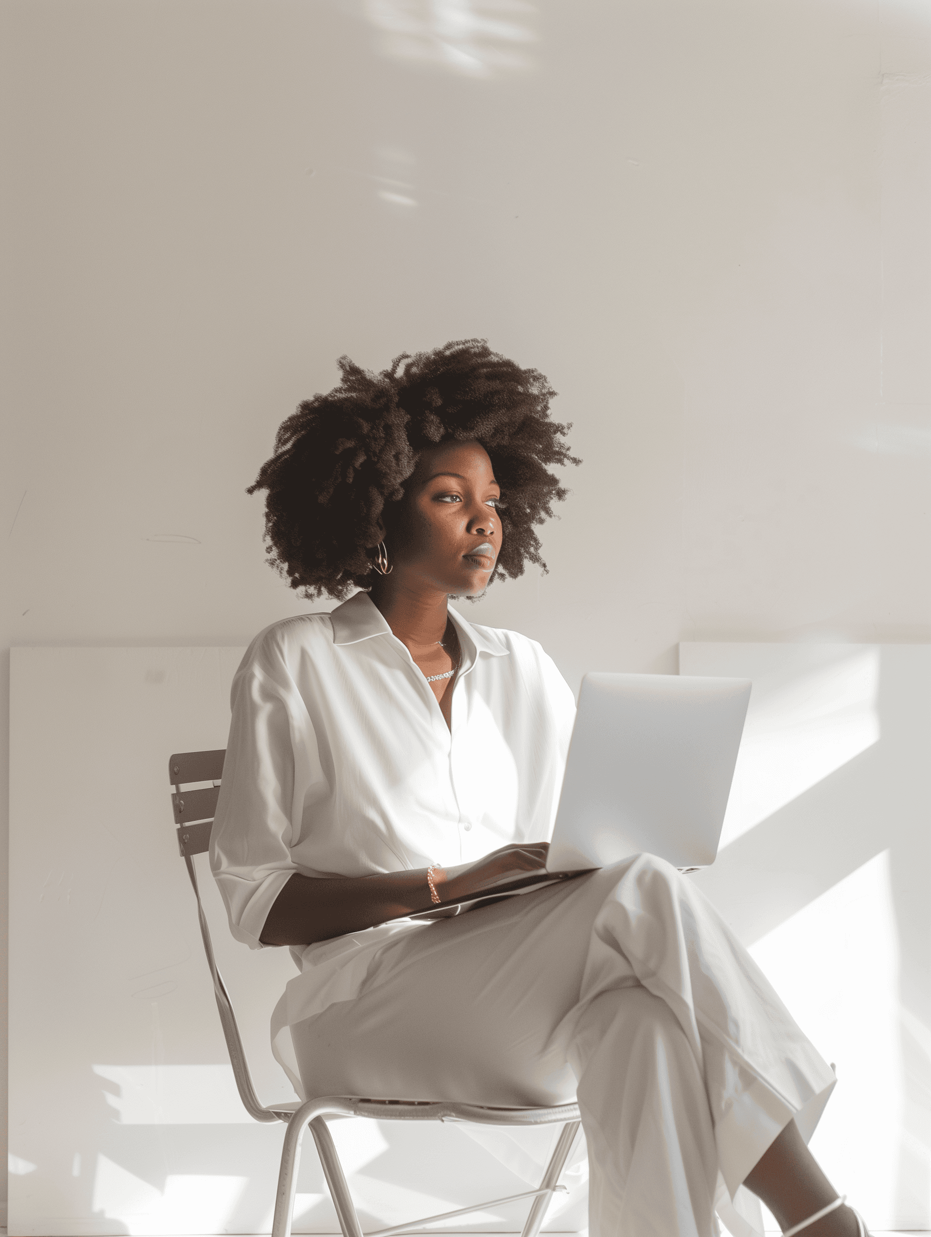 A young woman with natural afro hair sitting on a metal chair, wearing an all-white outfit, working on a laptop in a bright minimalist room.