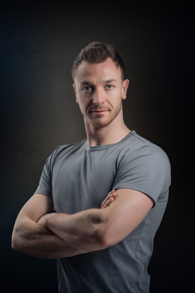 A smiling couple takes a selfie under a clear blue sky. The woman wears a red jacket, and the man, in a green top, holds a smartphone.