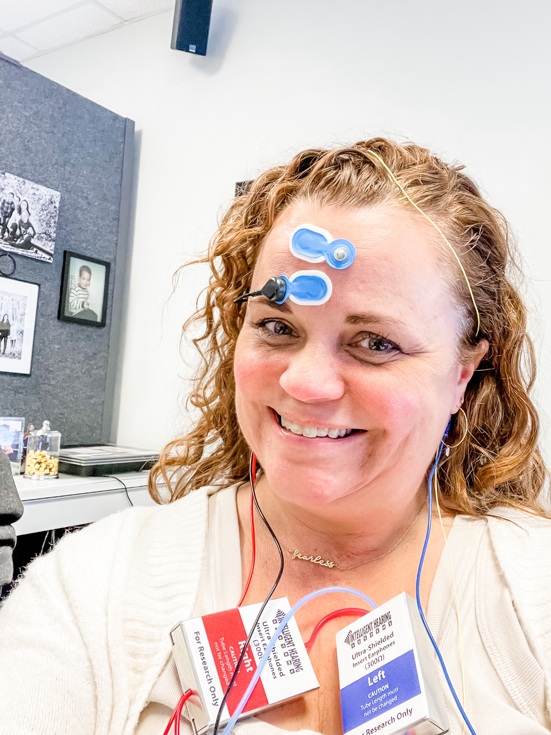 Dr Heidi Hill with curly hair is smiling in an office setting, wearing electrodes on her forehead, suggesting a brain or neural activity test.