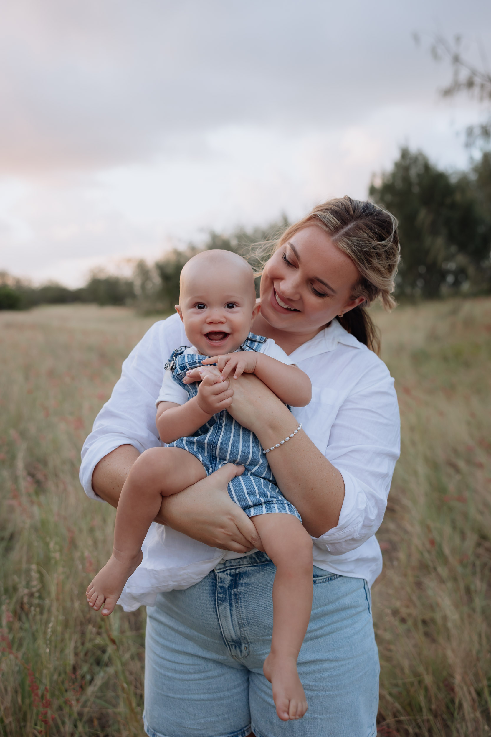 Mother holding her baby in tall golden grass during a warm outdoor motherhood session in Mackay Queensland