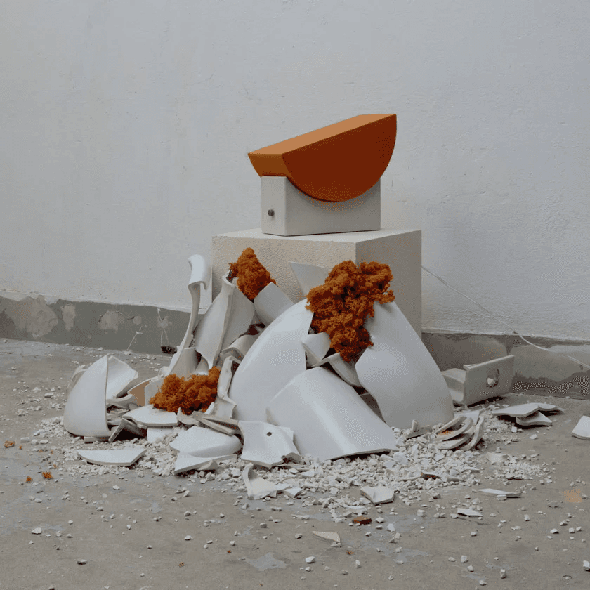 A modern art installation featuring a broken white ceramic sphere surrounded by rubble and orange mossy textures on a concrete floor against a plain white wall.