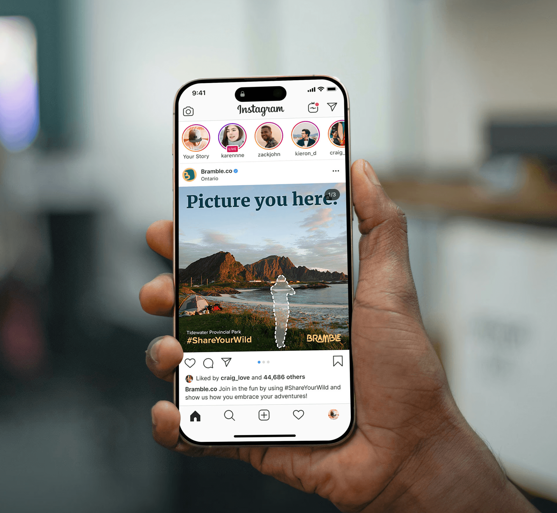 This image shows a person holding a smartphone that displays an Instagram post from the account Bramble.co. The post features a scenic photograph of a coastline with mountains and a tent at Tidewater Provincial Park, overlaid with the bold headline "Picture you here." and a dashed white silhouette of a person. A campaign hashtag, #ShareYourWild, and the Bramble logo are visible at the bottom of the image, while the caption encourages users to join in by sharing how they embrace their adventures. The phone's interface also shows various user story icons at the top and standard engagement features like likes and comments at the bottom.