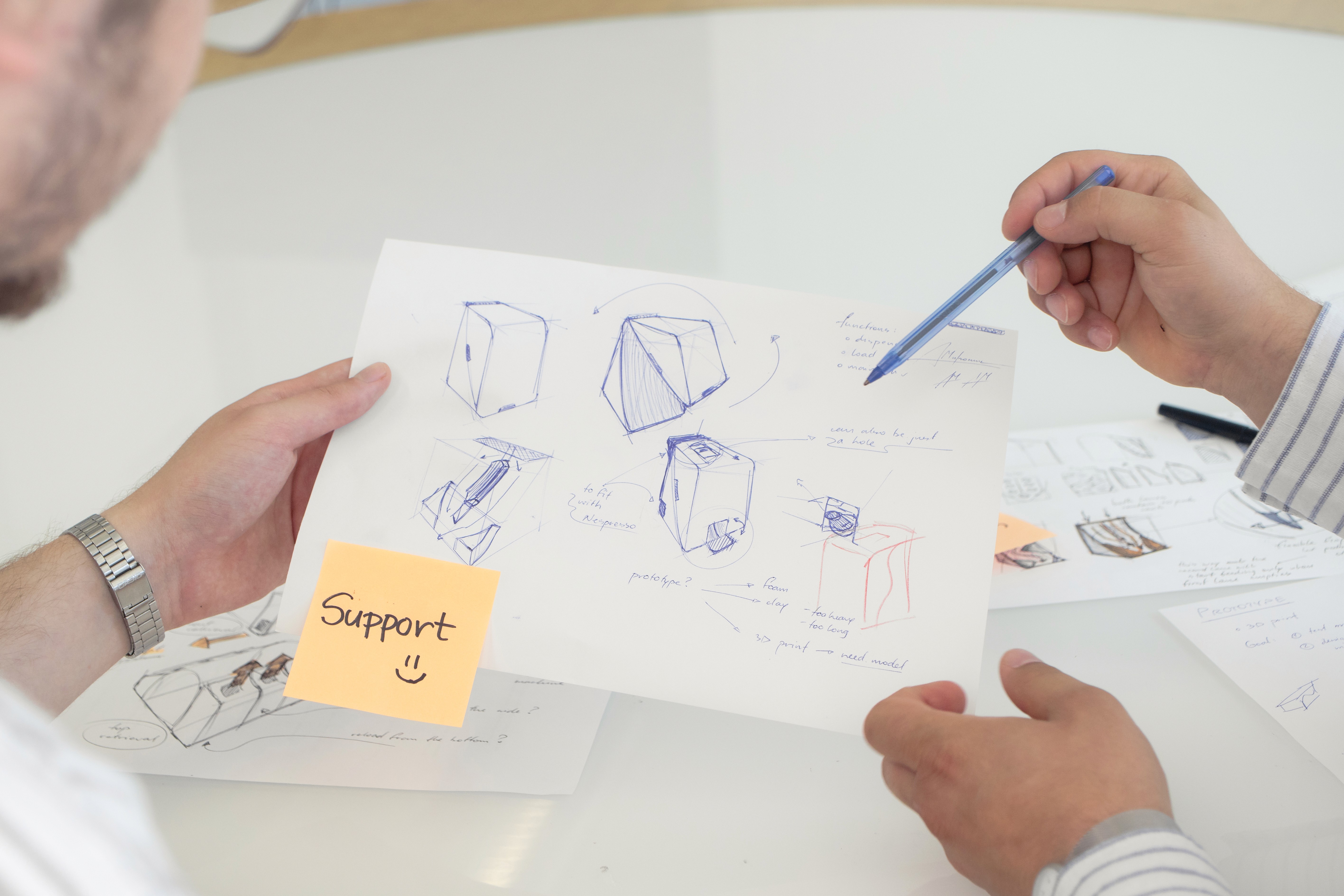 Two people collaborate over sketches of geometric designs on paper, one annotating with a blue pen. A note marked "Support" adds a positive touch.