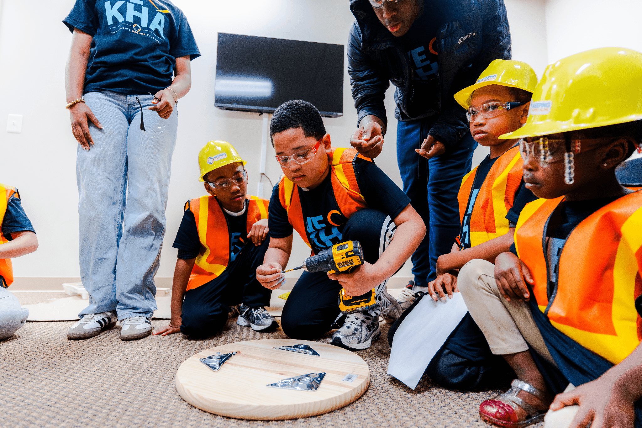 Youth wearing safety gear participate in a hands-on workshop using a power drill with adult supervision.