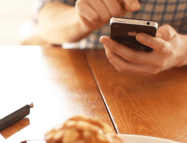 A person wearing a blue and white checkered shirt holds a black smartphone in their right hand, with their left hand positioned near the phone. A wooden table is visible beneath them. On the table, a dark-colored object resembling a cigarette rests to the left, and a blurred, light-colored object, possibly a pastry or cookie, is in the foreground. A white plate is partially visible in the bottom right corner of the image.