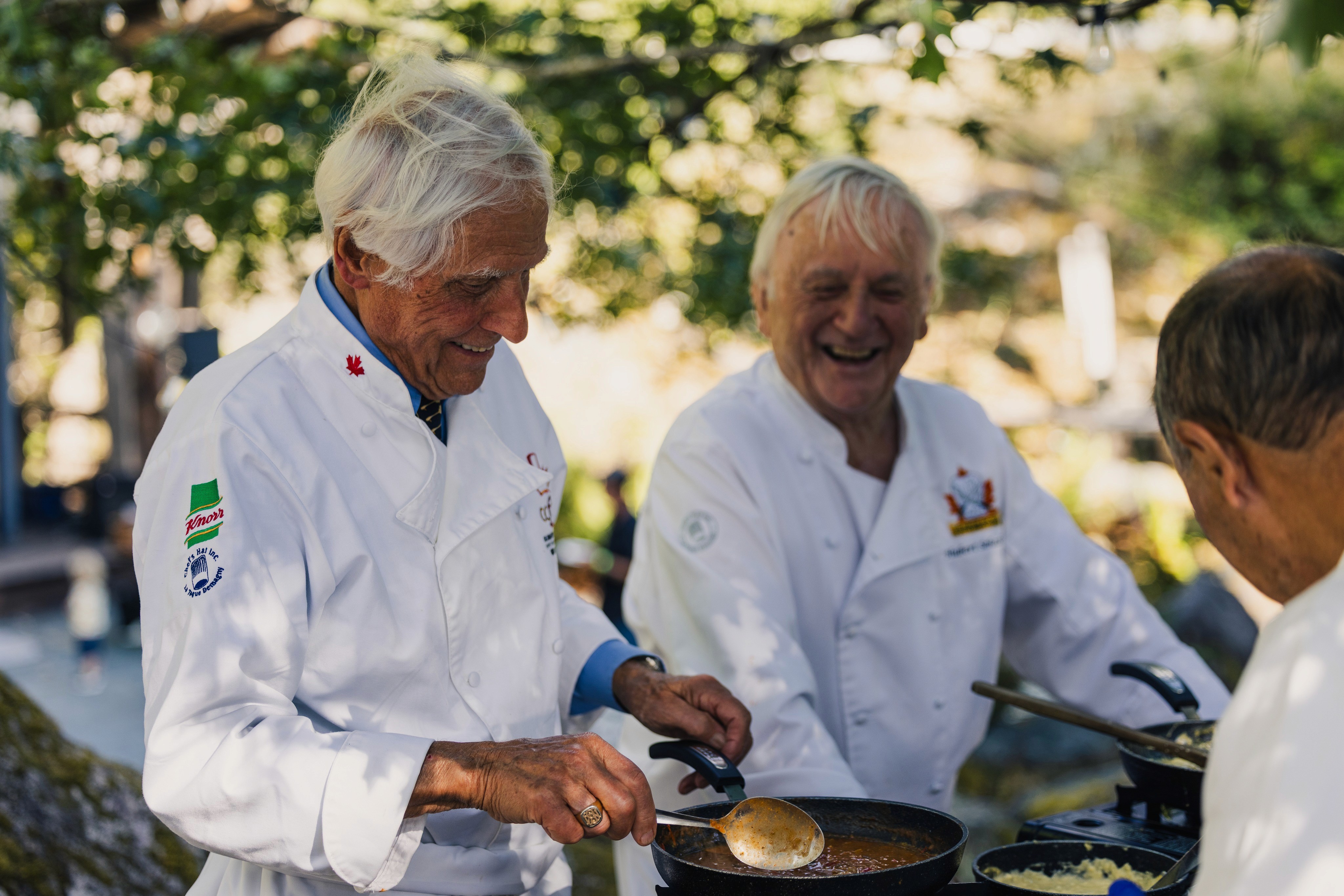 Two veteran chefs laughing while cooking a sauce outdoors