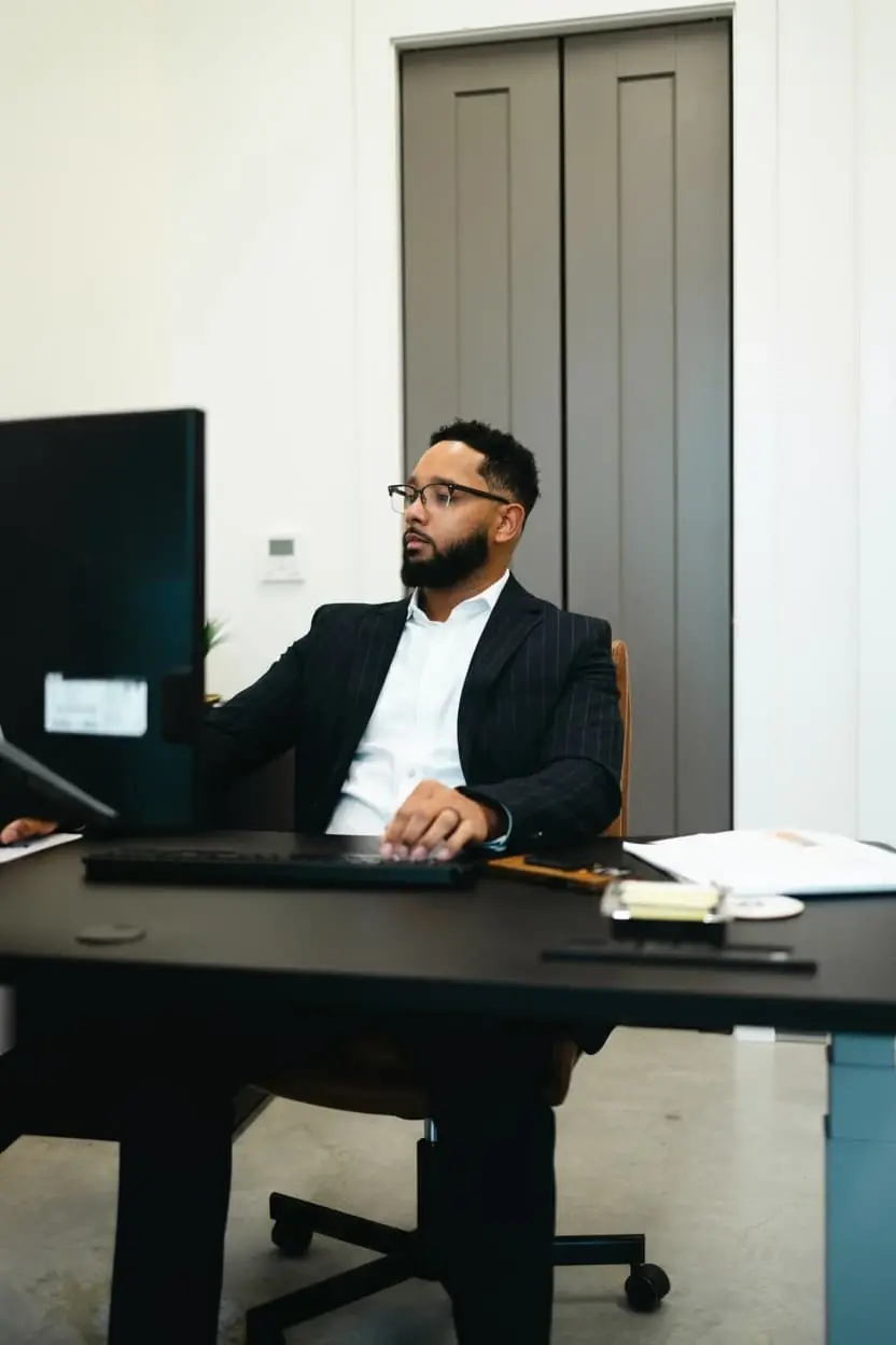 Chris Lewis works at his office desk wearing a dark suit and glasses, focused on his computer. The professional environment conveys trust, expertise, and dedication to helping clients secure smart investment loans through Chris Lewis Home Loans.