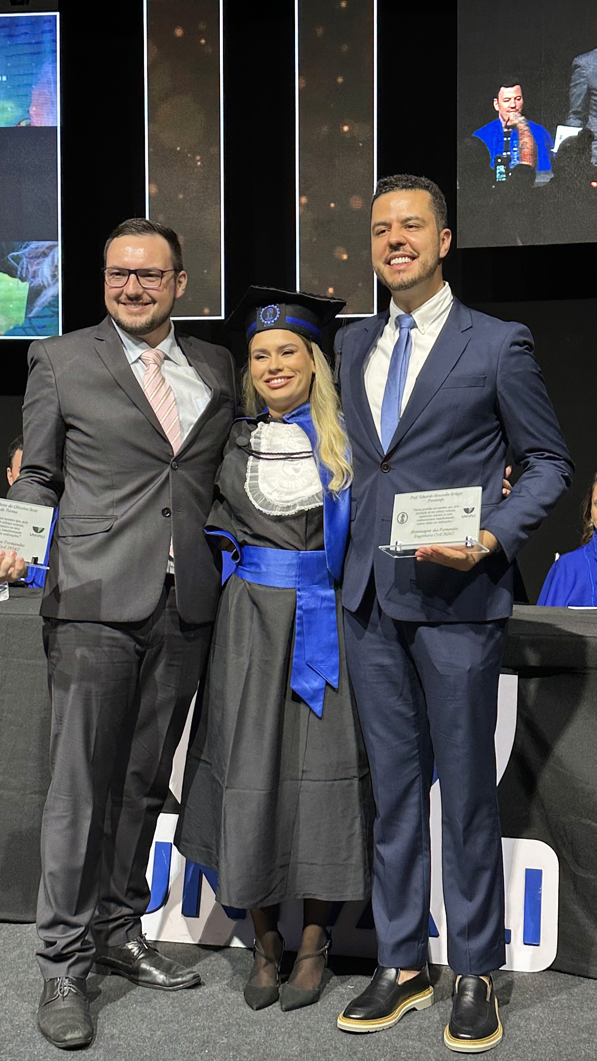 O professor Eduardo Alexandre Kruger e outro docente de terno segurando placas de homenagem acadêmica ao lado de formanda de Engenharia Civil da Univali.