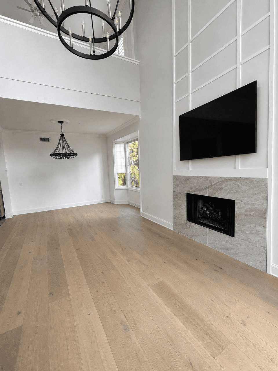 Renovated living room with light wood flooring, tiled fireplace, mounted television, and black chandeliers.