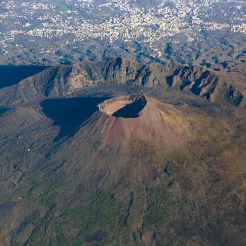 Aerial view of a large volcanic crater surrounded by rugged terrain and a distant cityscape in the background.