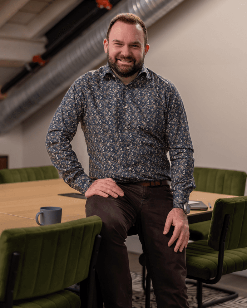 A smiling accountant, Alex Ataman, leans against a large wooden conference table surrounded by green chairs in a modern office with industrial ceiling ducts, while a coffee mug and notebook sit on the table.