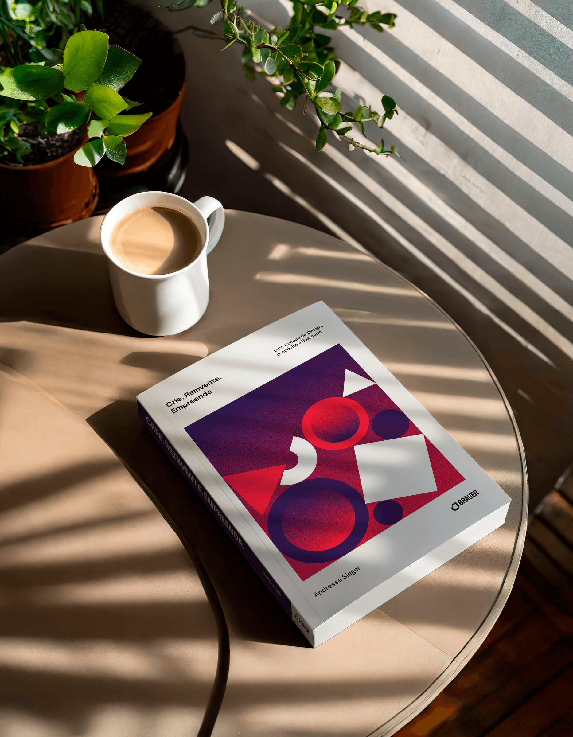 Livro "Crie. Reinvente. Empreenda." de Andressa Siegel sobre uma mesa de madeira, ao lado de uma xícara de café. A luz do sol atravessa as persianas, criando sombras suaves. Ao fundo, plantas verdes em vasos adicionam um toque natural ao ambiente