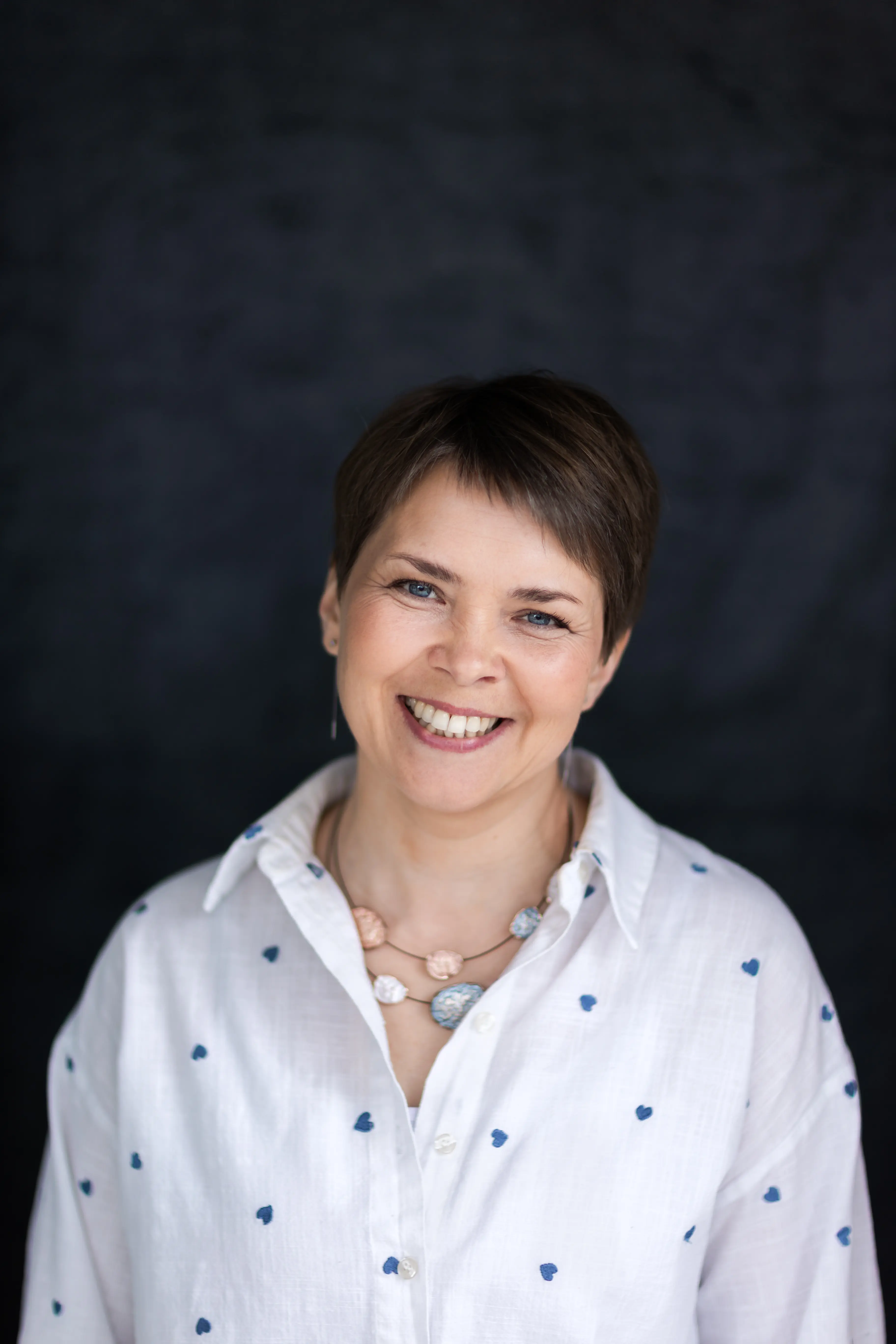 Portret kobiety o krótkich włosach uśmiechającej się na niebieskim tle.