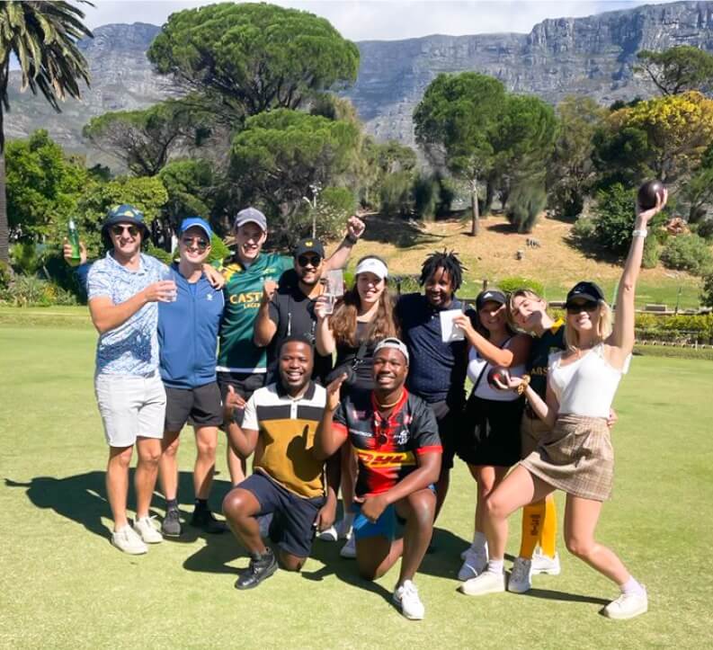 Outdoor team photo; diverse group cheering on a sunny day with a scenic mountain backdrop.