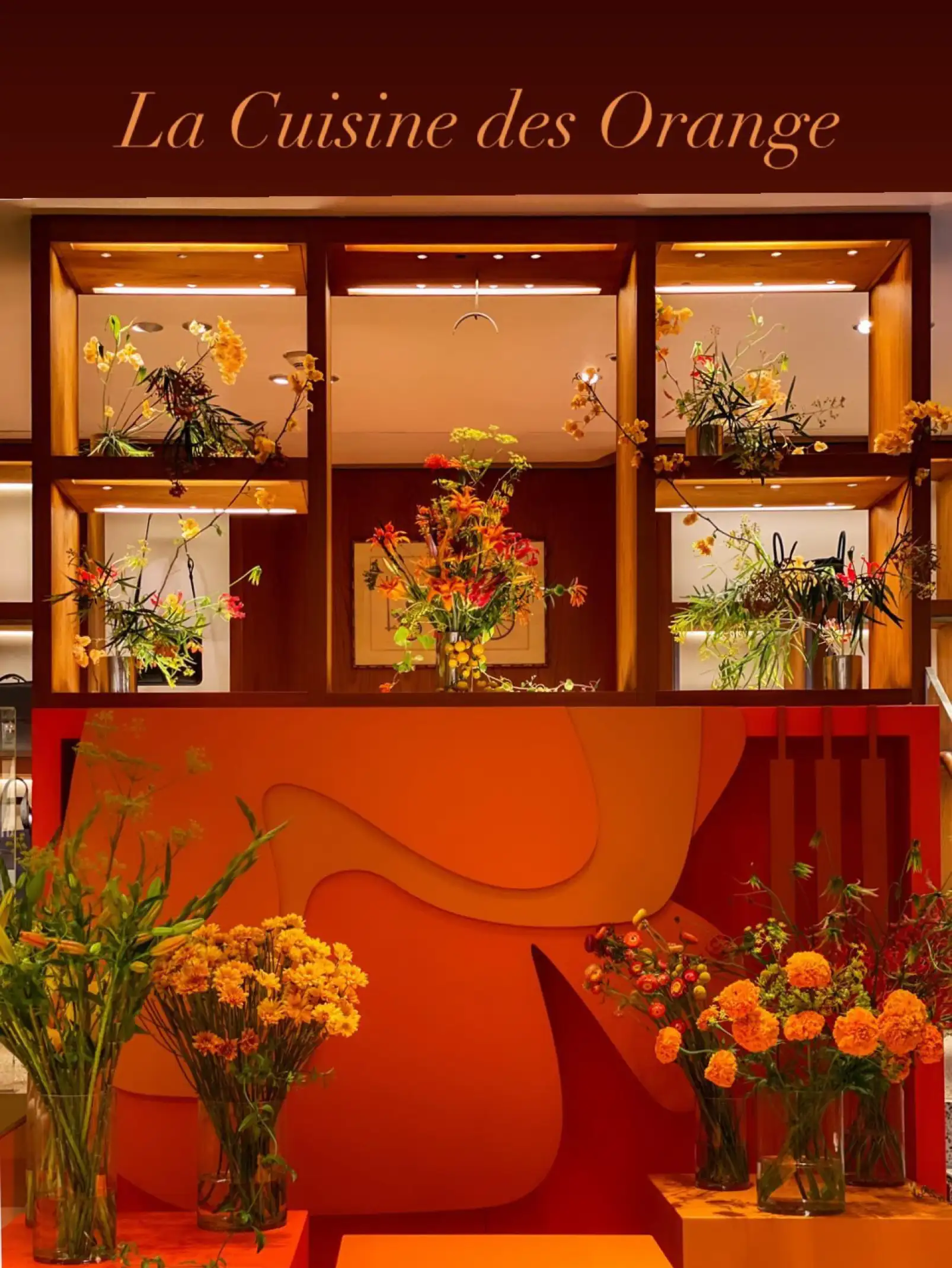 Instalación floral titulada La Cuisine des Orange, con arreglos de flores en tonos naranjas, rojos y amarillos distribuidos sobre pedestales y estantes de madera. El fondo presenta paneles escultóricos en degradé cálido, creando una atmósfera elegante y contemporánea que celebra la estética del color naranja.