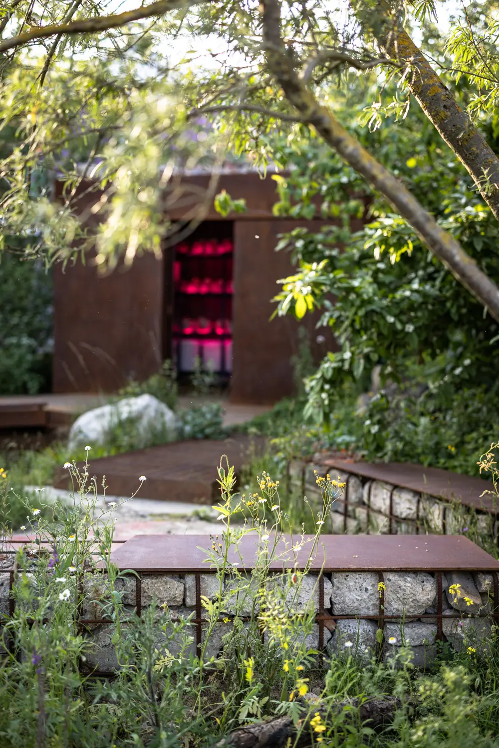 A serene garden scene featuring a brick building with a red door surrounded by lush greenery and a wooden path.