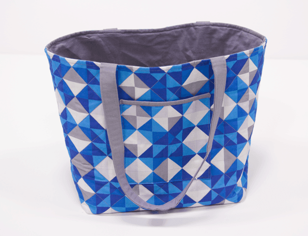 A blue and white geometric patterned tote bag with gray handles rests on a white surface. The bag features a pocket on the front and visible stitching along its edges. The interior lining is gray.