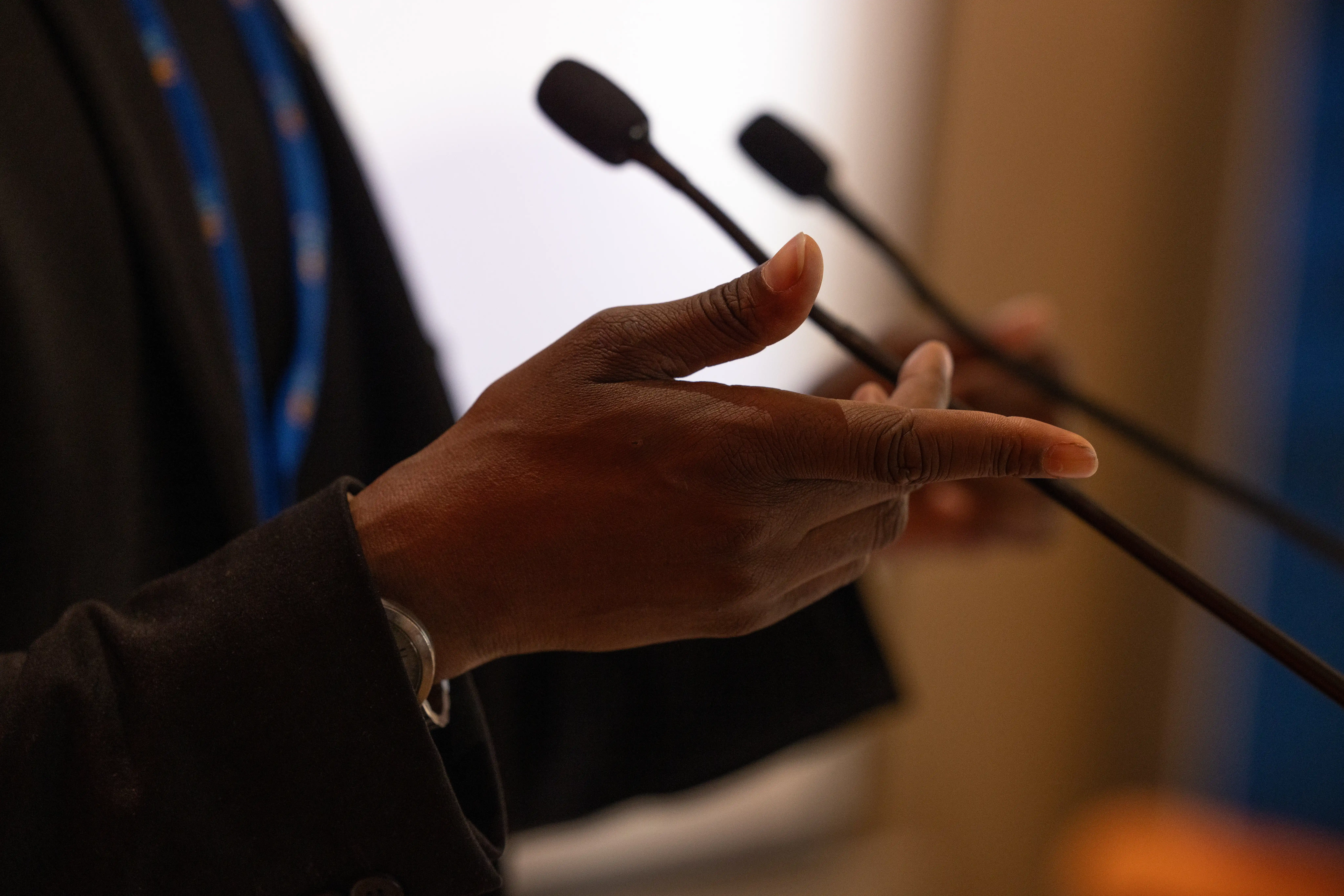 Close-up of a woman's hands wearing a lawyer's robe, speaking into a microphone, indicating she is giving a speech or engaging in a discussion.