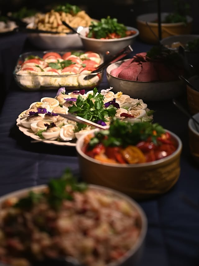 A long buffet table set for a meal, displaying multiple platters and bowls of food including pasta salad, tomatoes and mozzarella, sliced roast beef, and other prepared dishes.