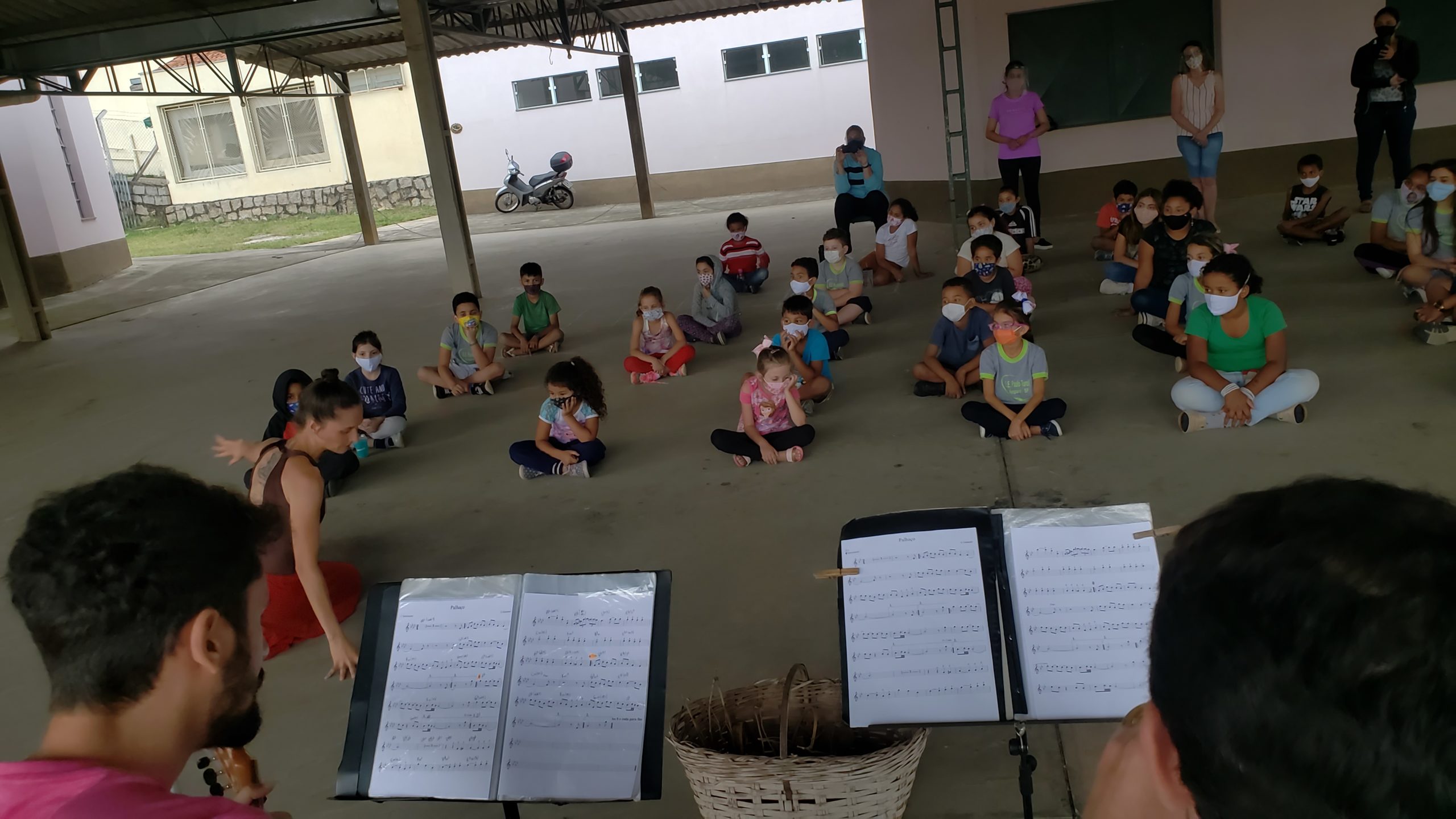 Alunos de máscara sentados no chão do pátio coberto assistem à bailarina, com partituras em primeiro plano