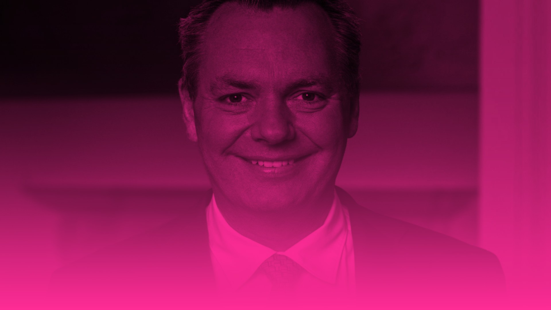 Das Foto zeigt ein eng herangezoomtes Porträt eines lächelnden Mannes mittleren Alters, der direkt in die Kamera blickt. Er hat kurzes, dunkles Haar, das an den Schläfen leicht ergraut ist, und trägt ein helles Hemd mit einer dezent gemusterten Krawatte unter einem dunklen Sakko. Wie bei den vorangegangenen Bildern dieser Serie liegt über dem gesamten Foto ein intensiver magentafarbener (pinker) Farbfilter. Der untere Bildrand verläuft fließend in eine durchgehende, leuchtend pinke Farbfläche, die etwa das untere Drittel des Bildes einnimmt. Der Hintergrund ist sehr dunkel und zeigt keine erkennbaren Details.