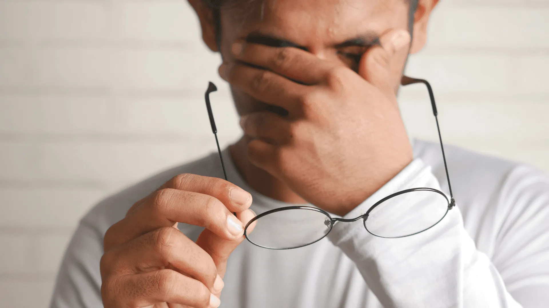 A man holds glasses and rubs his head over his eyes as though in pain