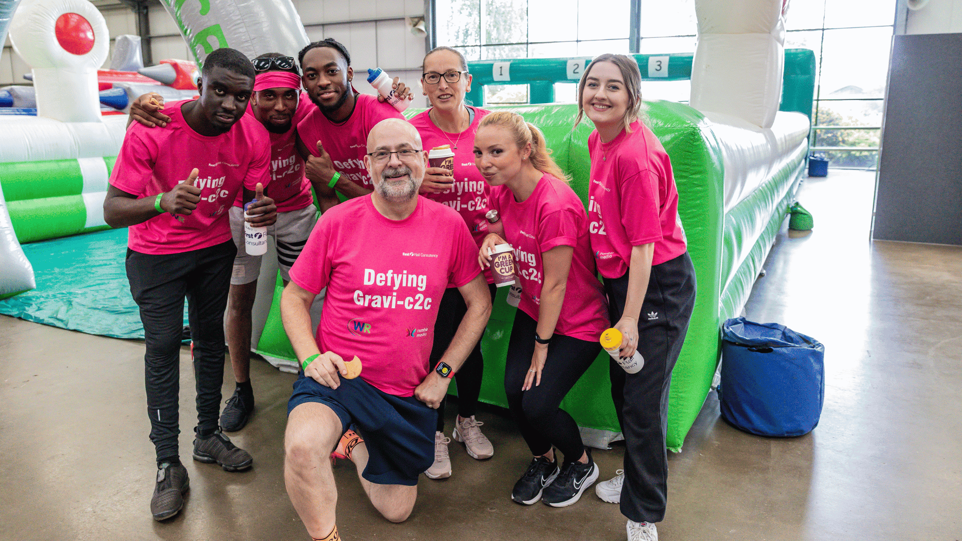 Outdoor team-building event with participants wearing matching blue shirts, taking part in inflatable obstacle courses and games, smiling and cheering, with the words 'ENTER A TEAM' in large bold white and green text across the image.