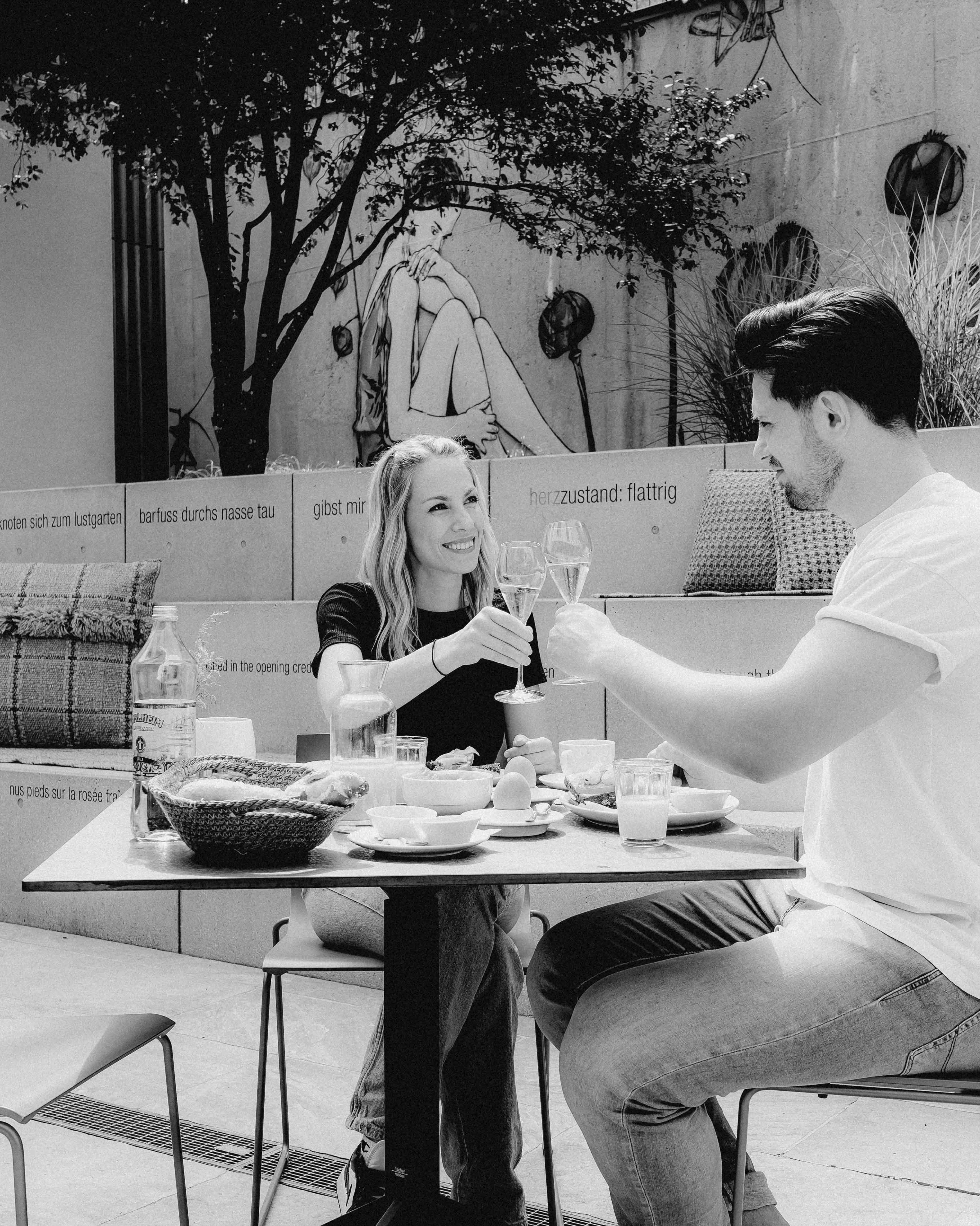 A couple enjoys a meal at an outdoor table, surrounded by trees, in a black-and-white photograph.