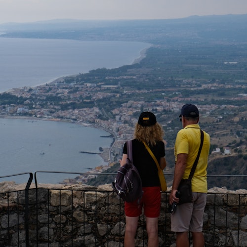 Deux personnes portant des chapeaux et des sacs se tiennent à une balustrade, surplombant une ville côtière et un paysage vaste.