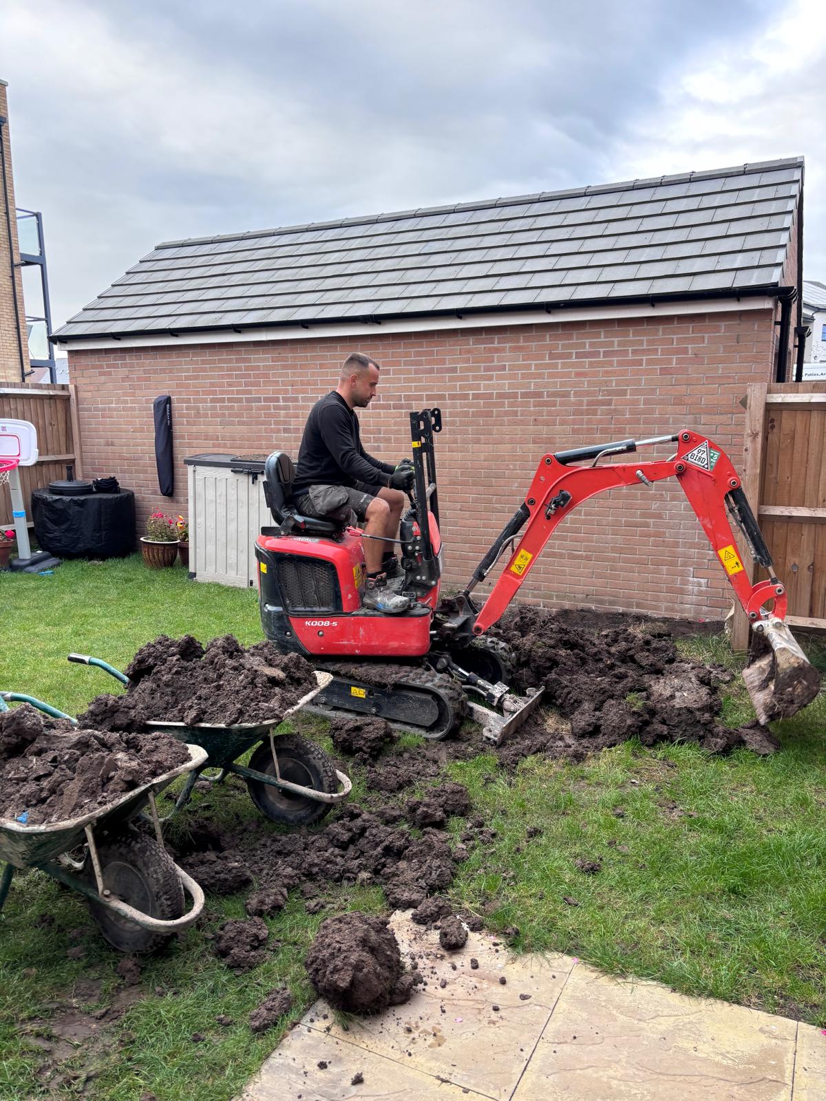 Bartec team member operating a red mini digger in a garden, with excavated soil and wheelbarrows beside a brick garage.