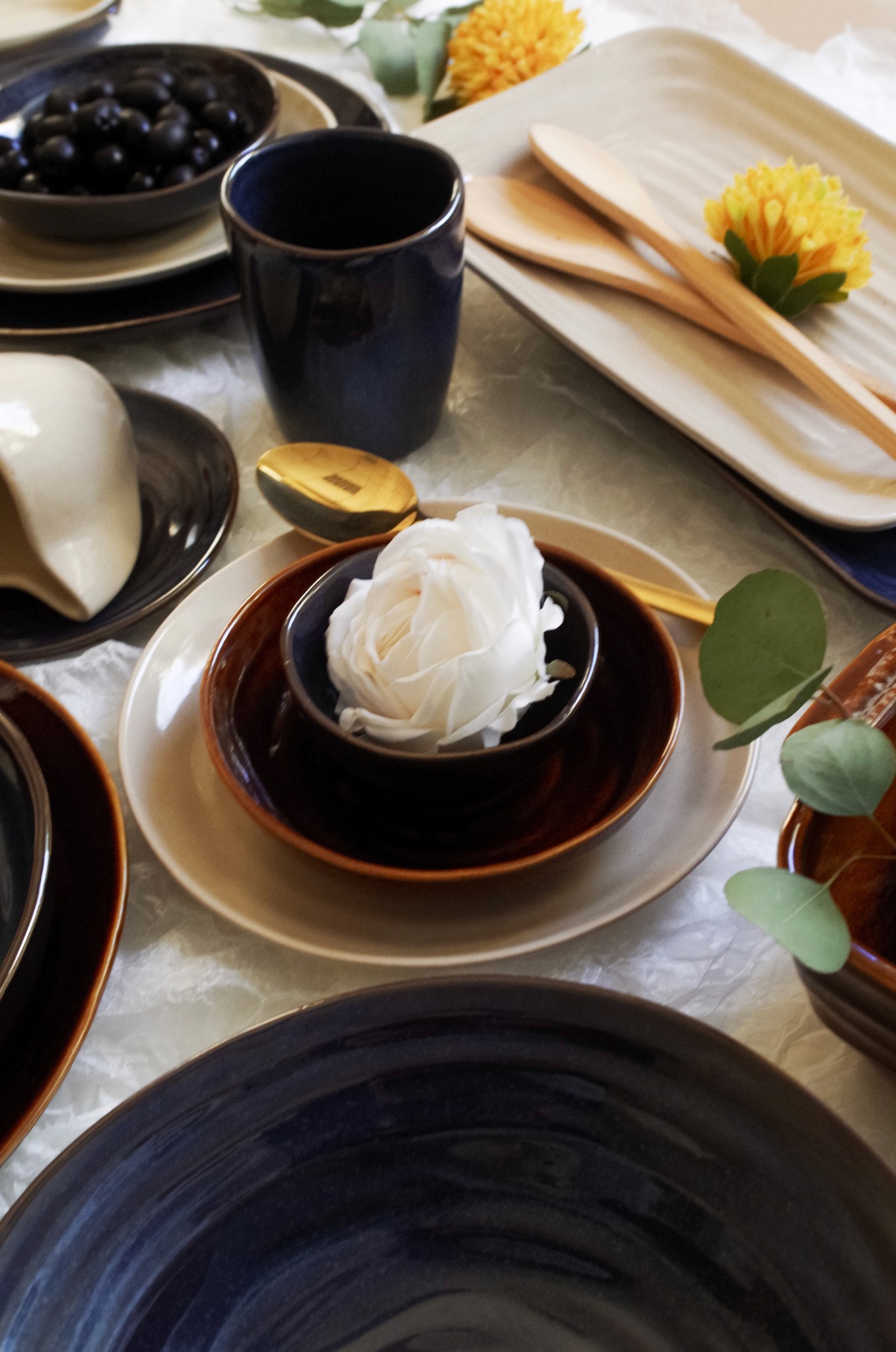 Détail d'une table dressée avec bol en céramique brun-bleu nuit contenant une rose blanche, posé sur assiette beige, avec couverts dorés et fleurs jaunes