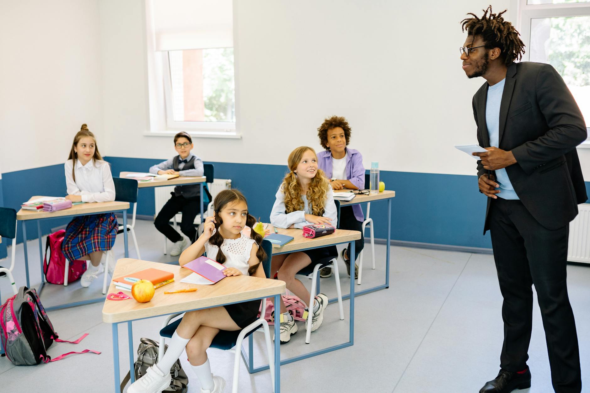 A smiling teacher points to a colorful leadership chart while elementary students raise their hands in a classroom.