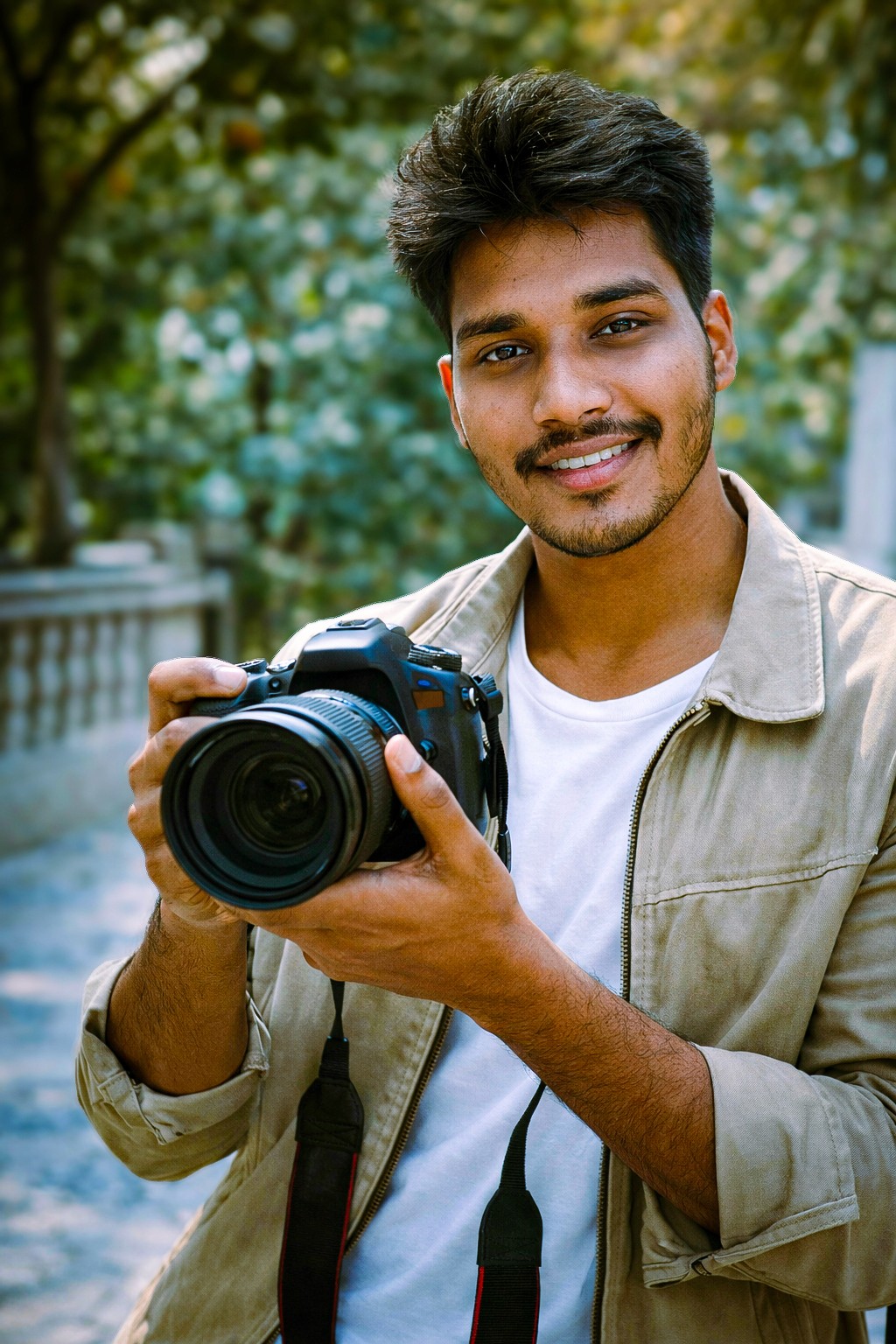 A man with a beard and sunglasses holding a camera with a long lens, wearing a jacket and backpack, looking at the camera.