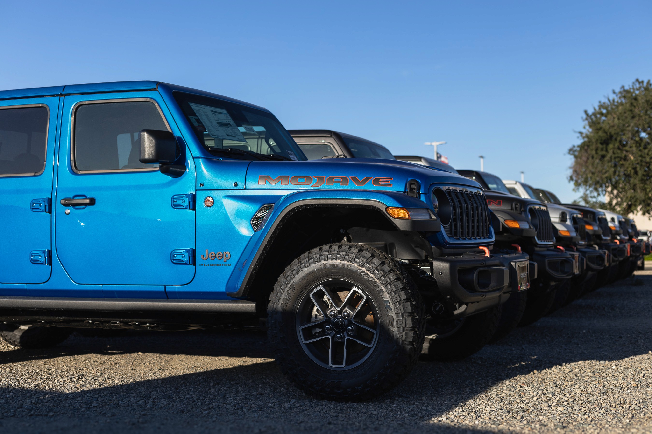 A line of rugged SUVs is parked on a gravel lot under a clear blue sky, with the focus on a vibrant blue model featuring the word "Mojave" on the hood.