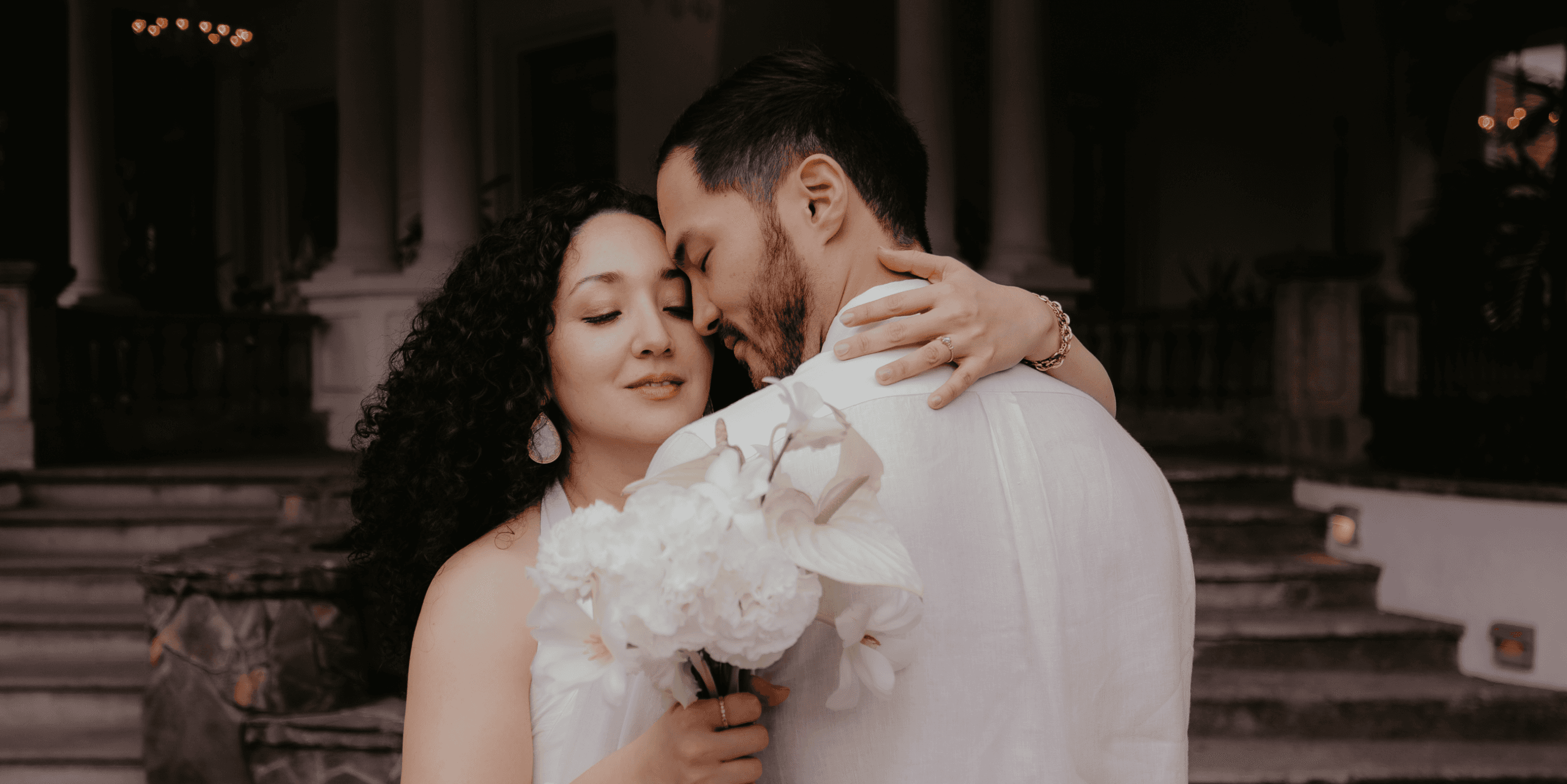A bride and groom hold hands tenderly, the bride's bouquet of dried flowers hanging low. They stand in a softly lit, elegant interior, conveying romance.