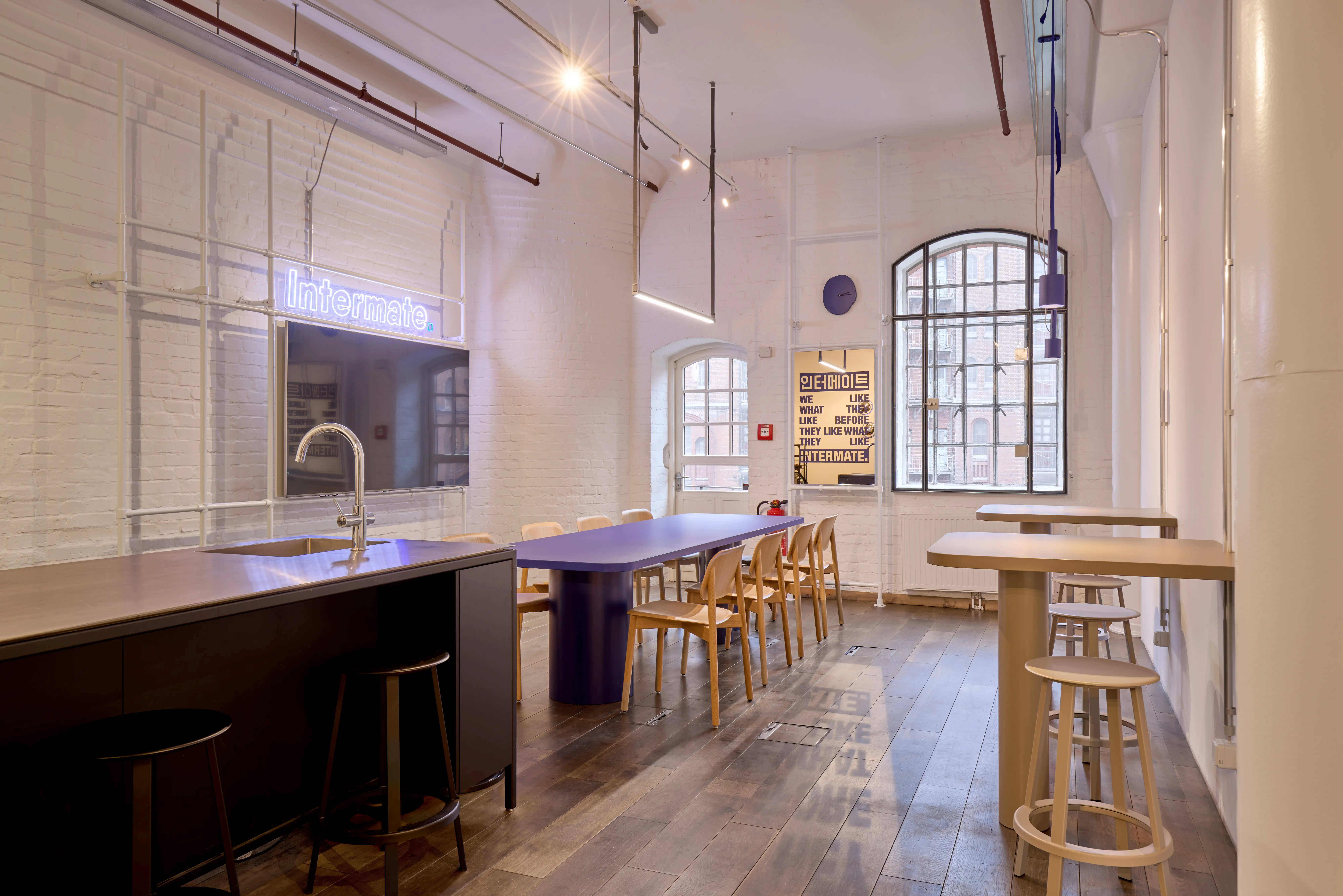 Kitchen and dining area at INTERMATE OFFICE HAMBURG with a dark kitchen island, a purple dining table with wooden chairs, beige standing tables with stools, a neon Intermate sign on white brick walls, and an arched factory window.