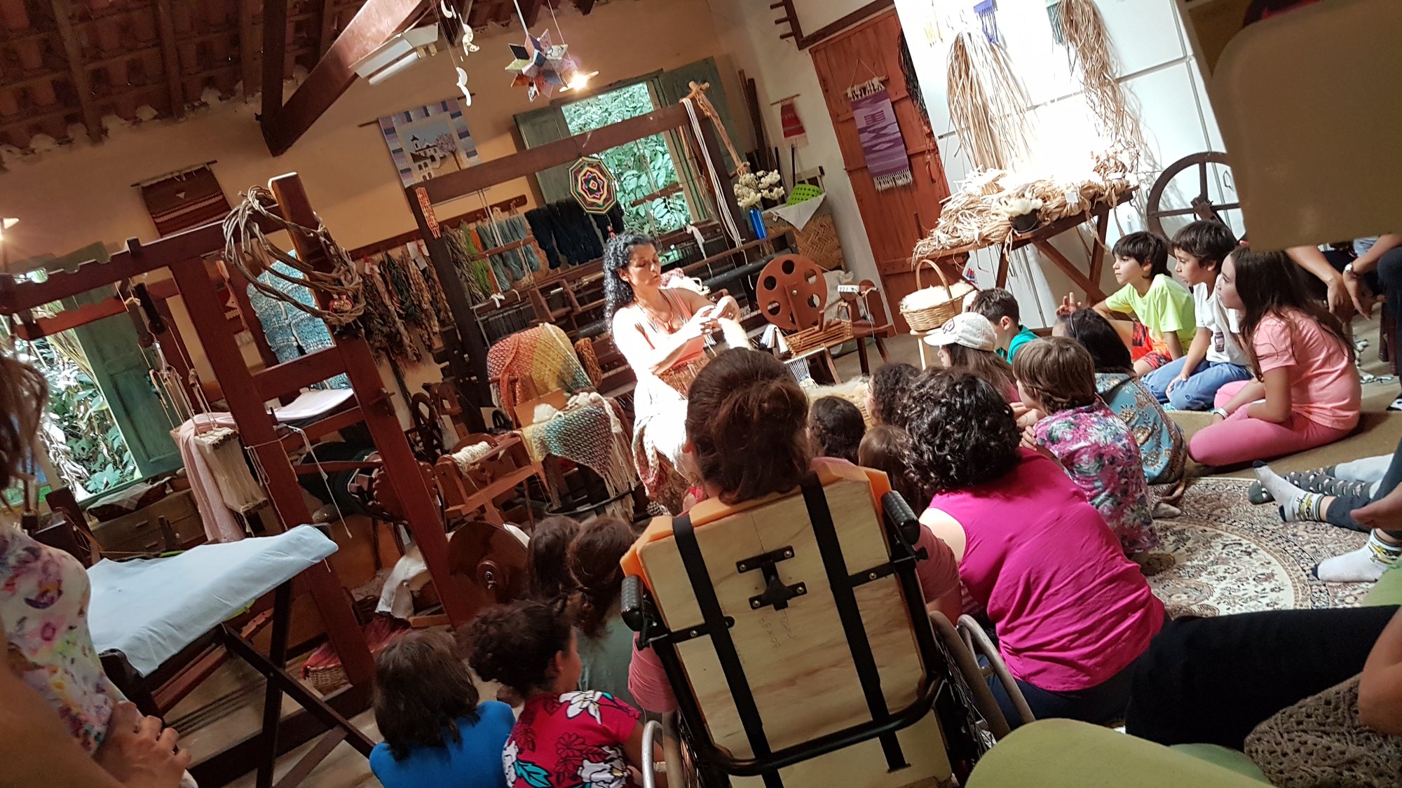 Crianças sentadas assistem a uma apresentação em sala de tecelagem com teares de madeira