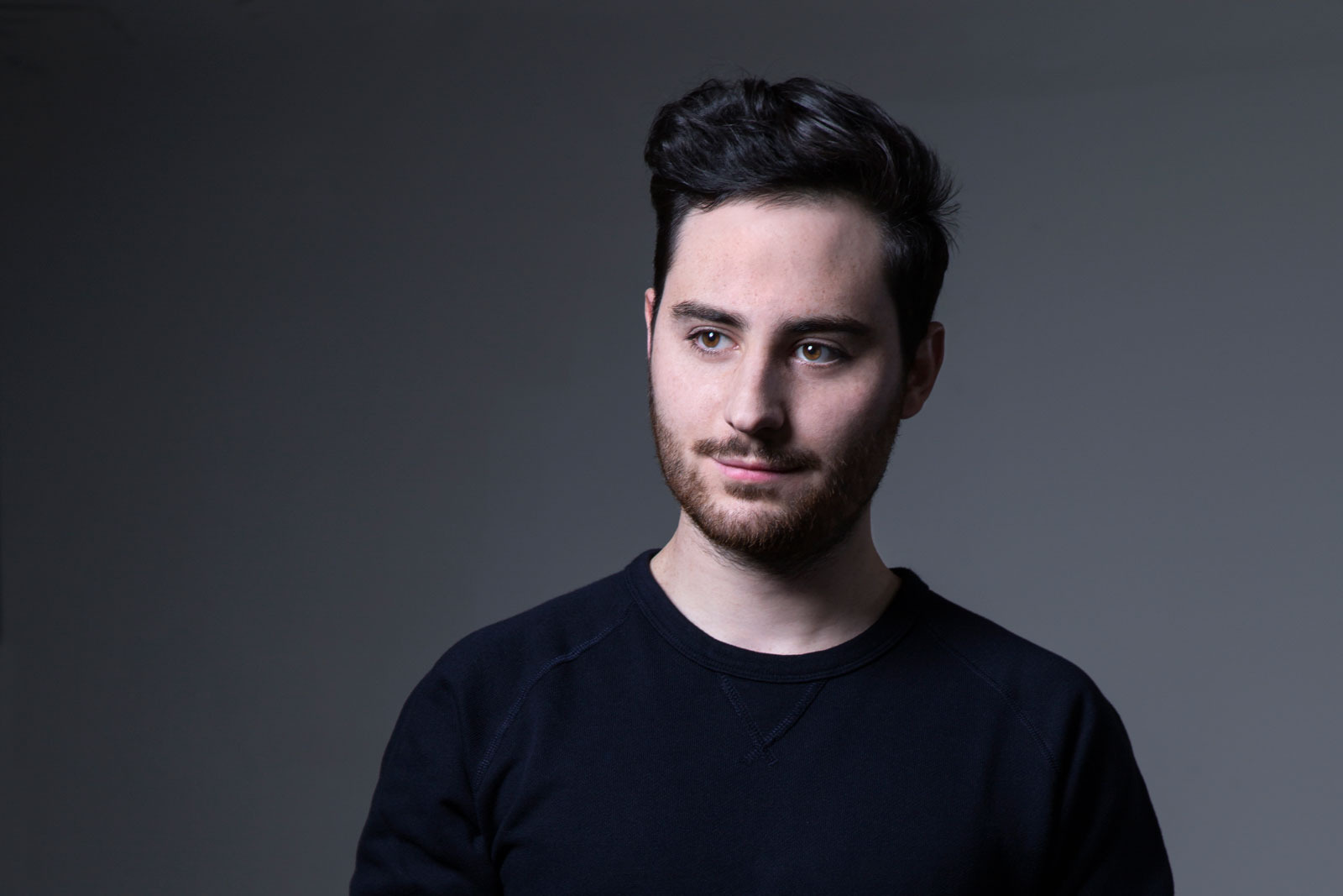 A young man with dark hair and a beard is captured in a head-and-shoulders portrait against a dark gray background. He wears a dark navy or black crew-neck shirt and looks directly at the camera with a neutral expression. His hair is styled in a slightly tousled, textured look, and his facial features are clear and distinct. The lighting is even and soft, highlighting his face without harsh shadows.
