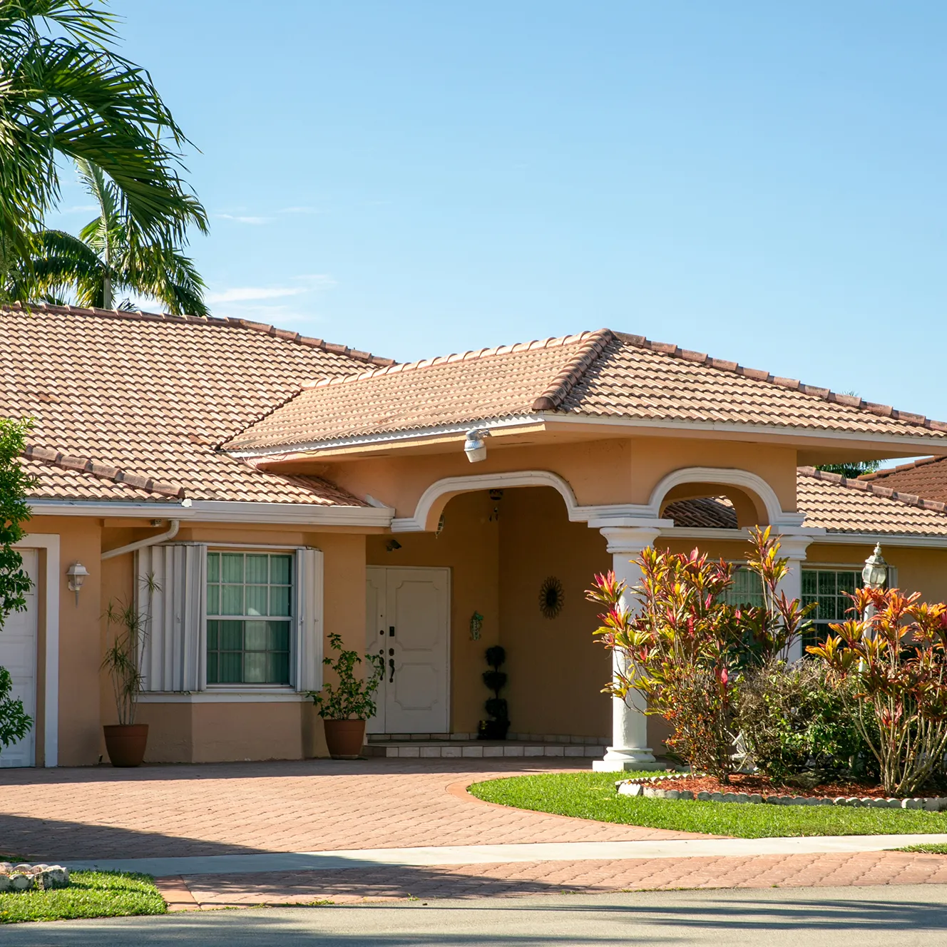 Single-story house with a terracotta-tiled roof, archway entrance, plants in the yard, and a paved driveway. Bright, sunny day, tropical vibe.