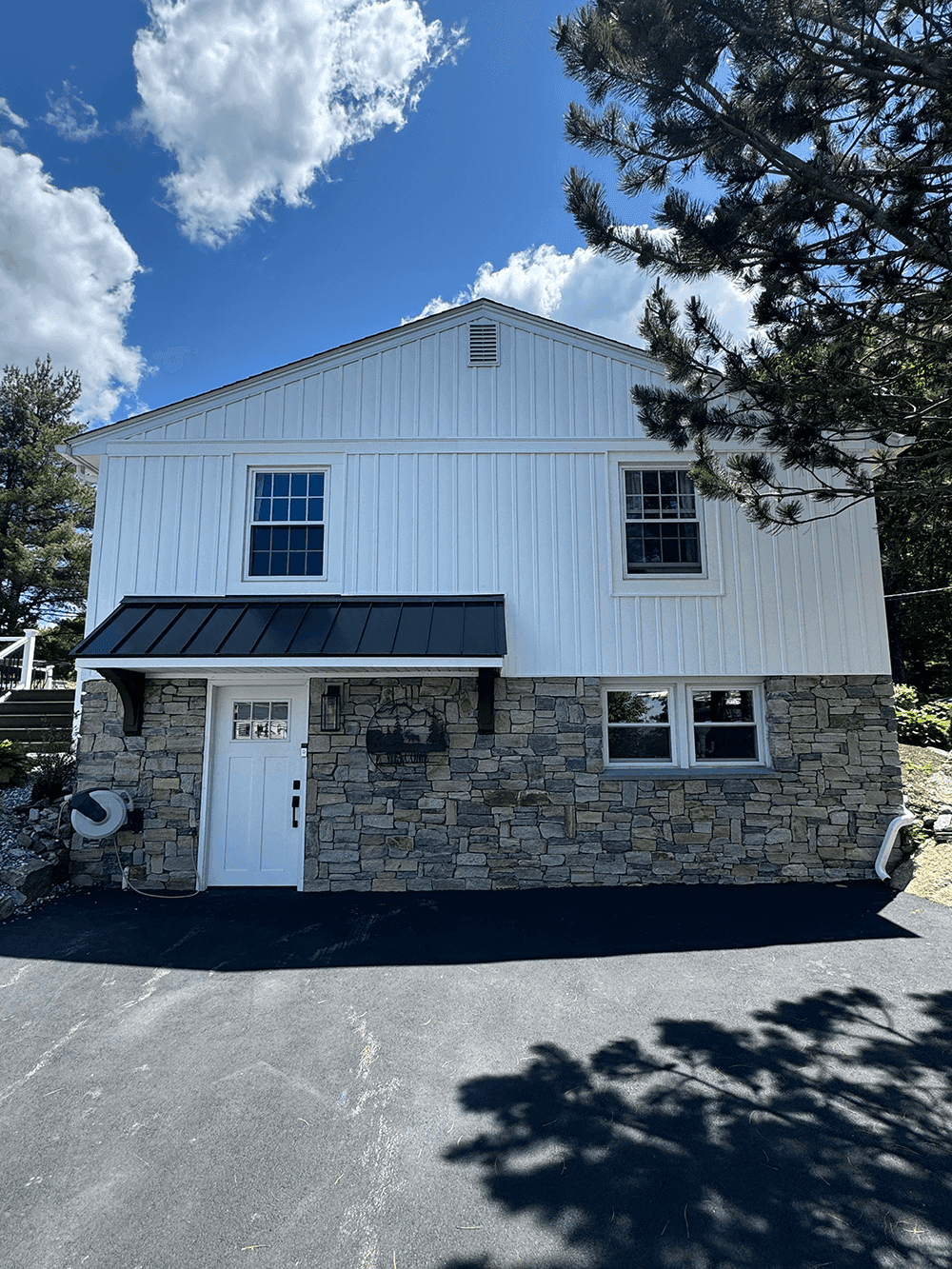 A house with a combination of white vertical panel siding on the upper section and stone veneer on the lower section. The entrance features a white door with a black metal awning supported by decorative brackets, set against the stone facade. The property is surrounded by trees, with a paved driveway in the foreground.