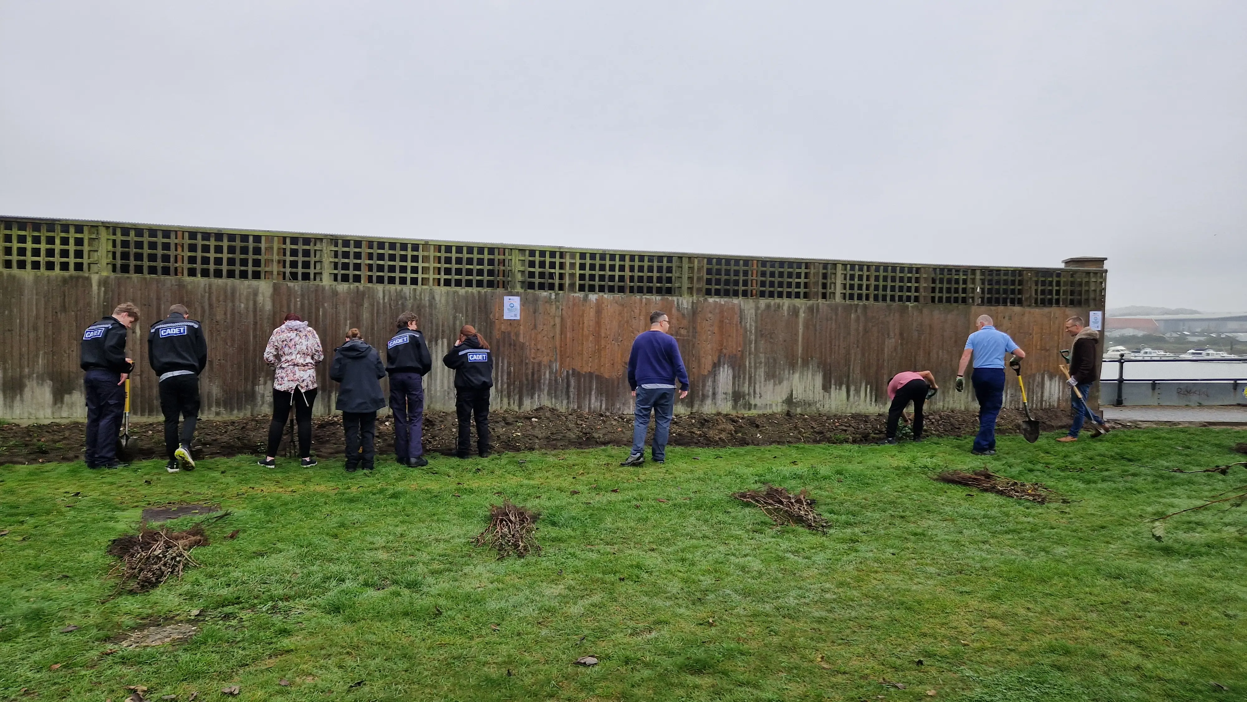 A group of people stands on grass near a brick wall in a cloudy outdoor setting.