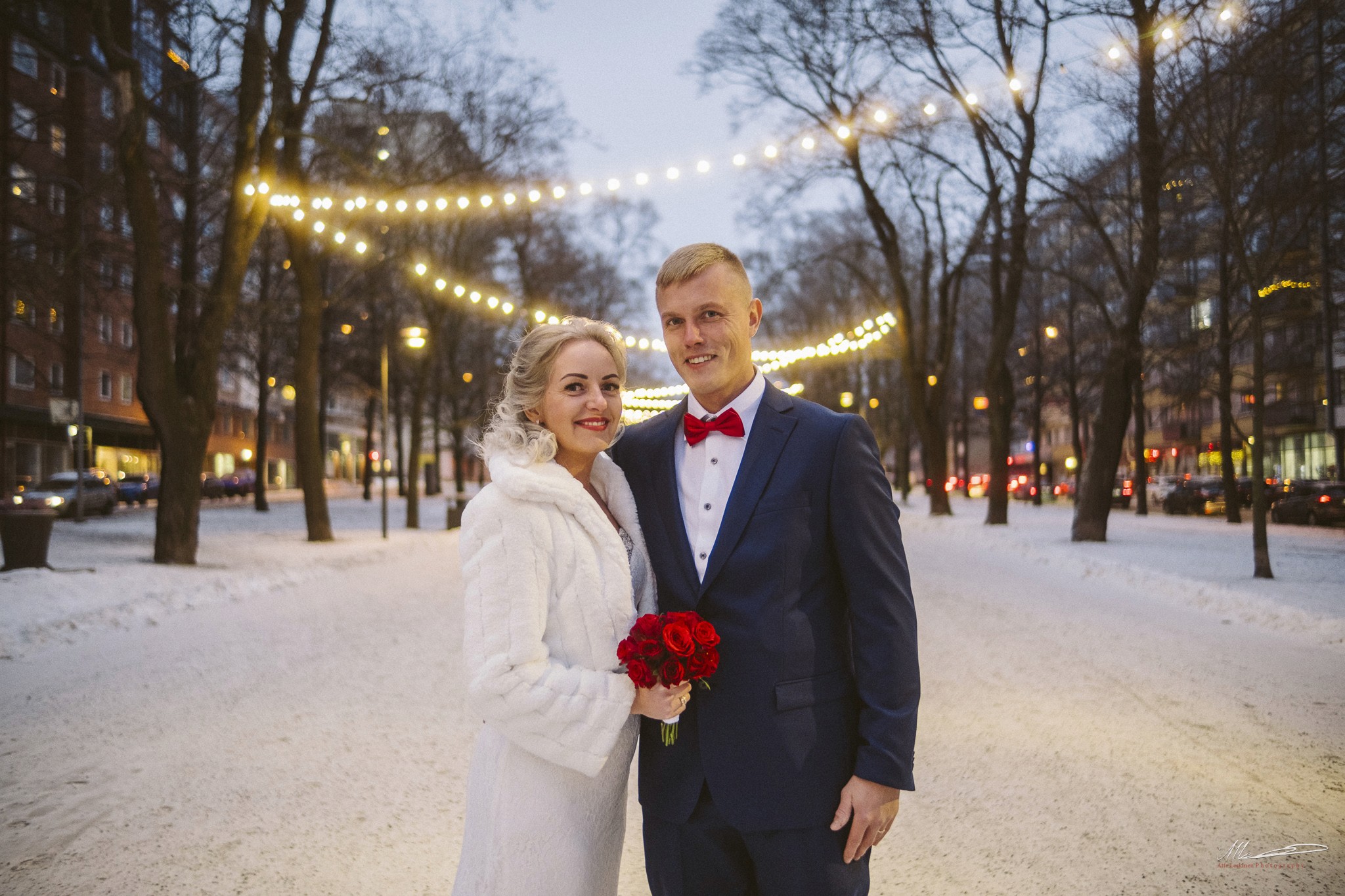 Talvihäät kaupungissa: hääpari lumisella puistokadulla valosarjojen alla, morsiamella punainen ruusukimppu