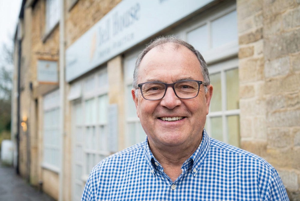 A man with glasses in a blue checkered shirt stands smiling in front of a stone building with large windows and a sign for Bell House Dental Practice.